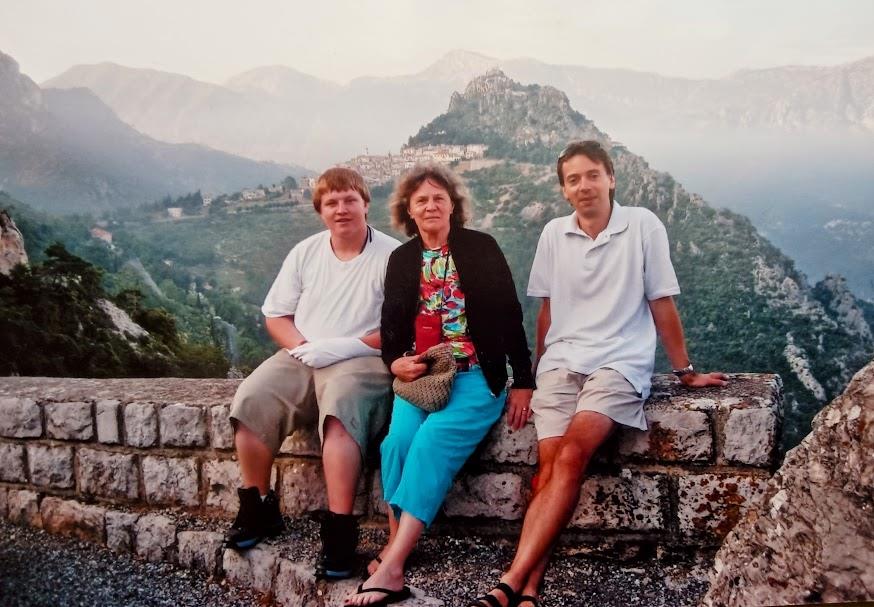 Three individuals relax on a stone wall with a breathtaking mountain backdrop during a warm evening.