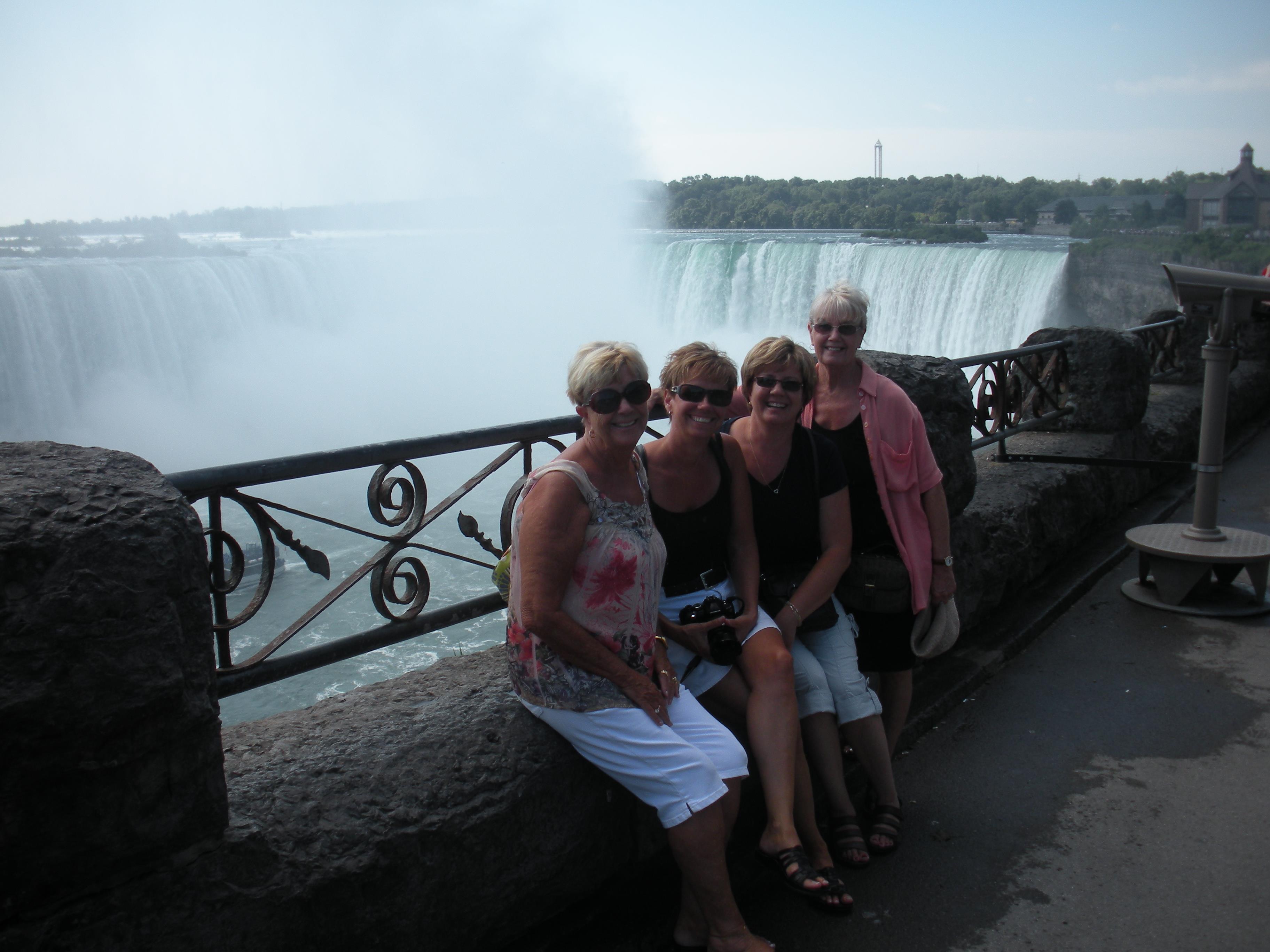 Four friends smile on a stone ledge at Niagara Falls, enjoying the summer sun.