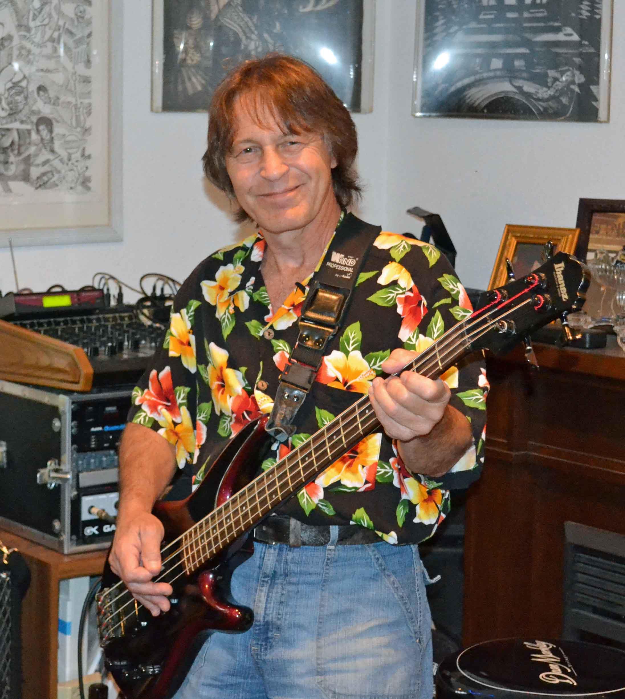 Musician plays electric bass while smiling and dressed in a colorful floral shirt at a party.