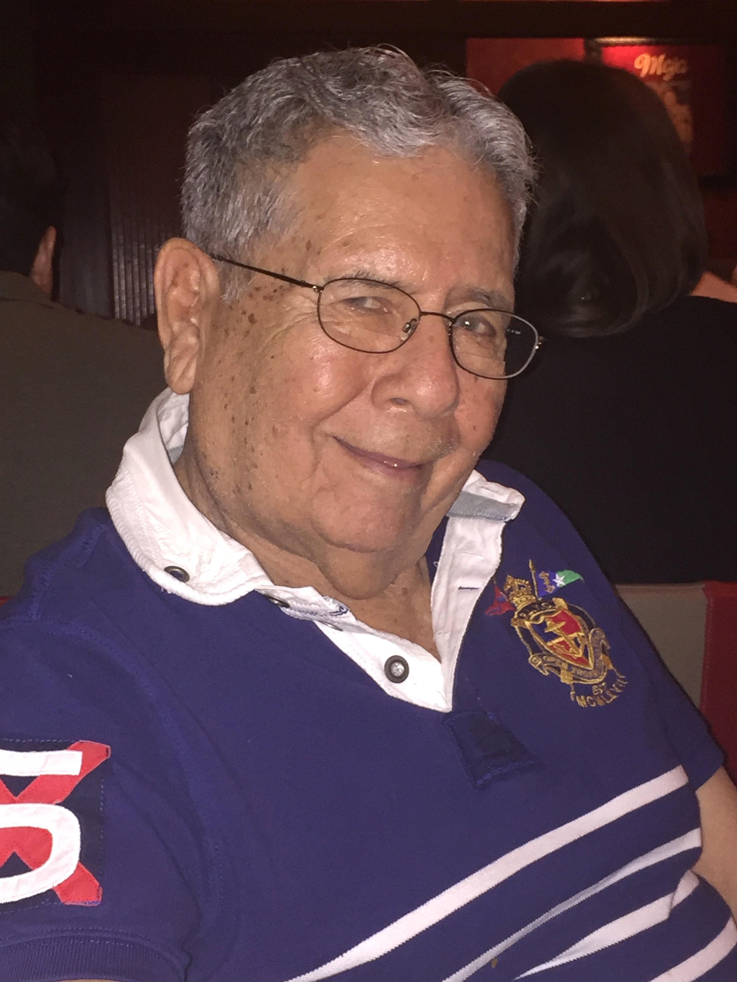An elderly man sits at a table, smiling kindly amidst a lively atmosphere during a night gathering.