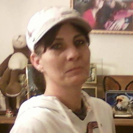 A woman with a neutral expression stands in a room filled with personal belongings and decorations.