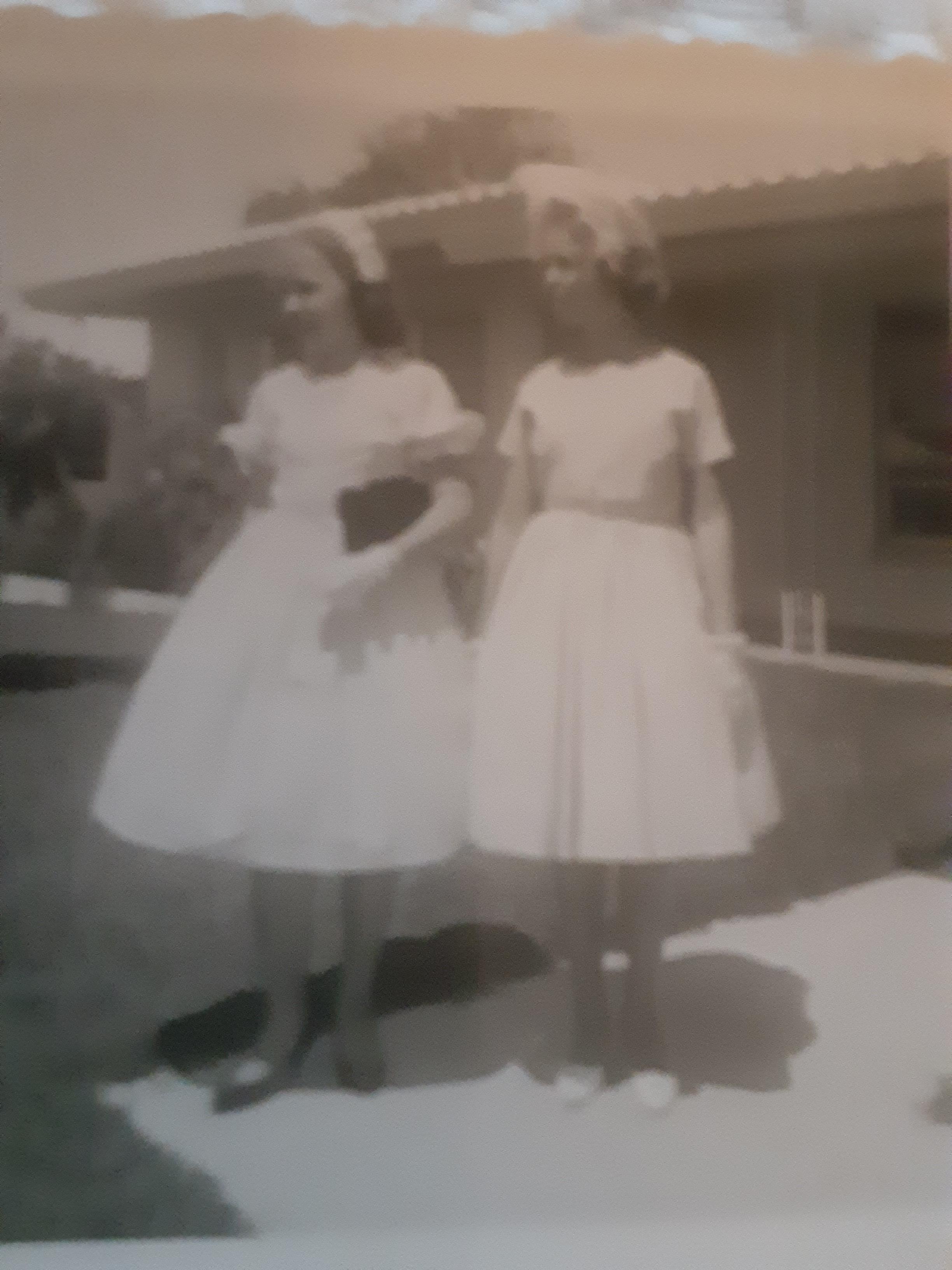 Two girls wear white dresses and smile together outside a house on a bright day.
