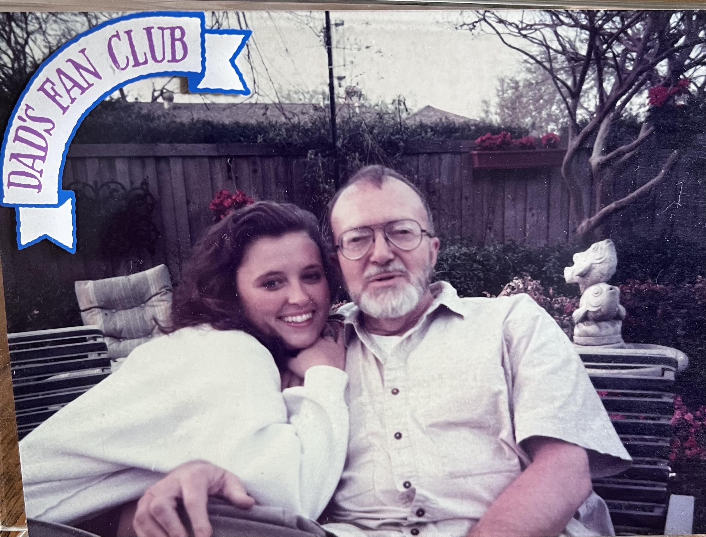 A man and his daughter smile while sitting outdoors in a garden setting filled with flowers.