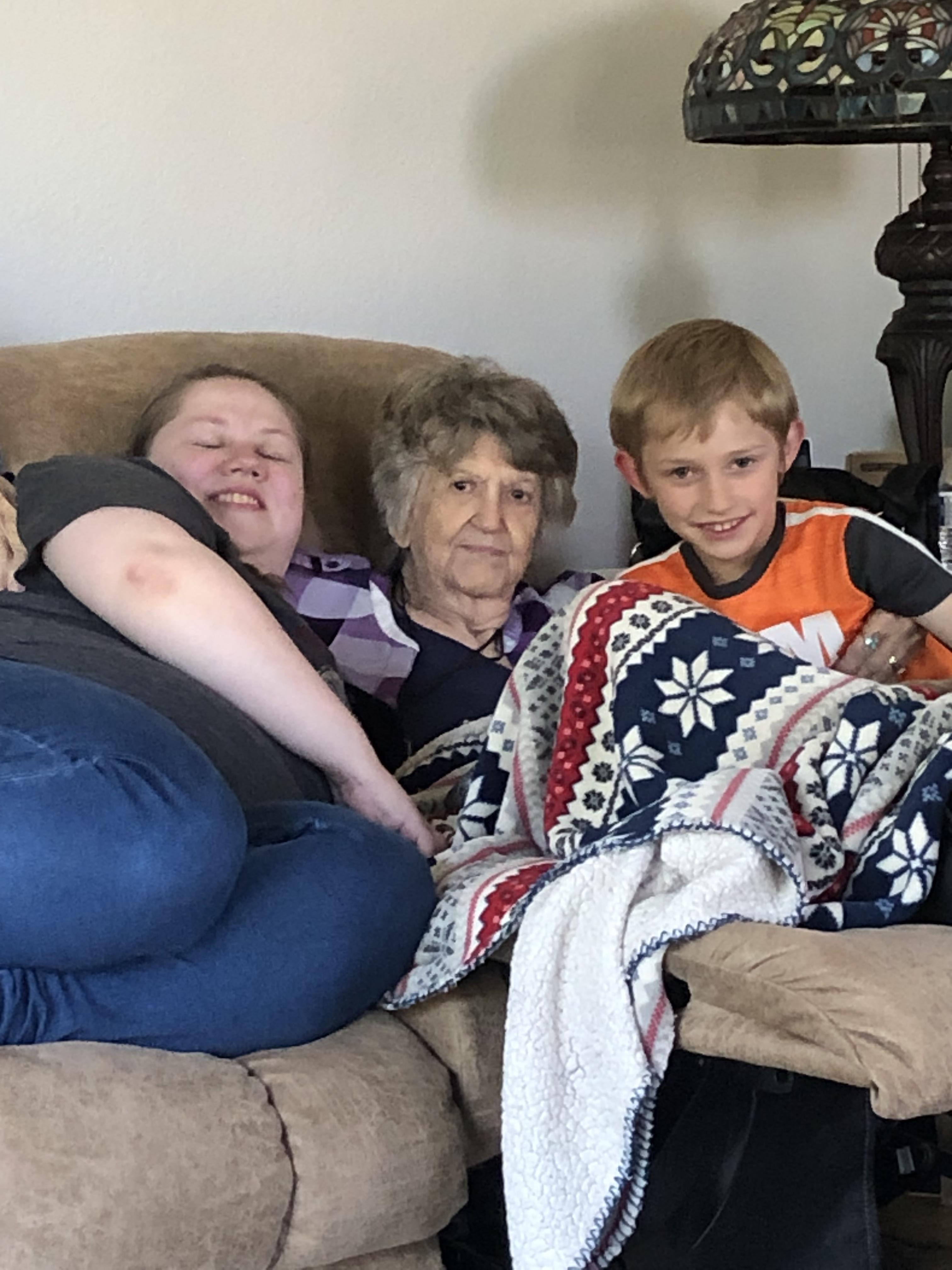 Three family members relax together on a couch, wrapped in a blanket, sharing smiles and warmth.