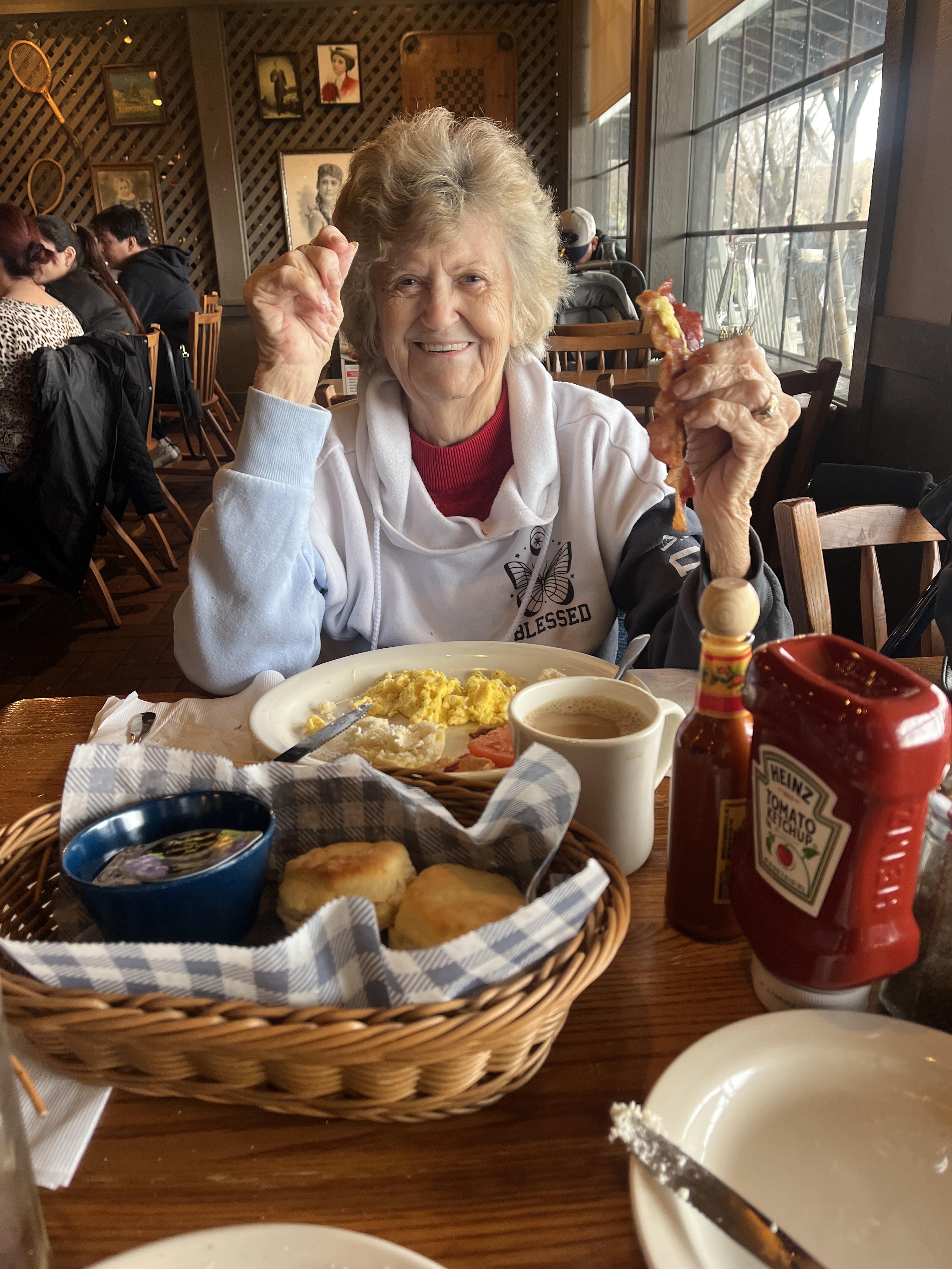 Elderly woman smiles while holding food in a diner, surrounded by breakfast dishes and coffee.