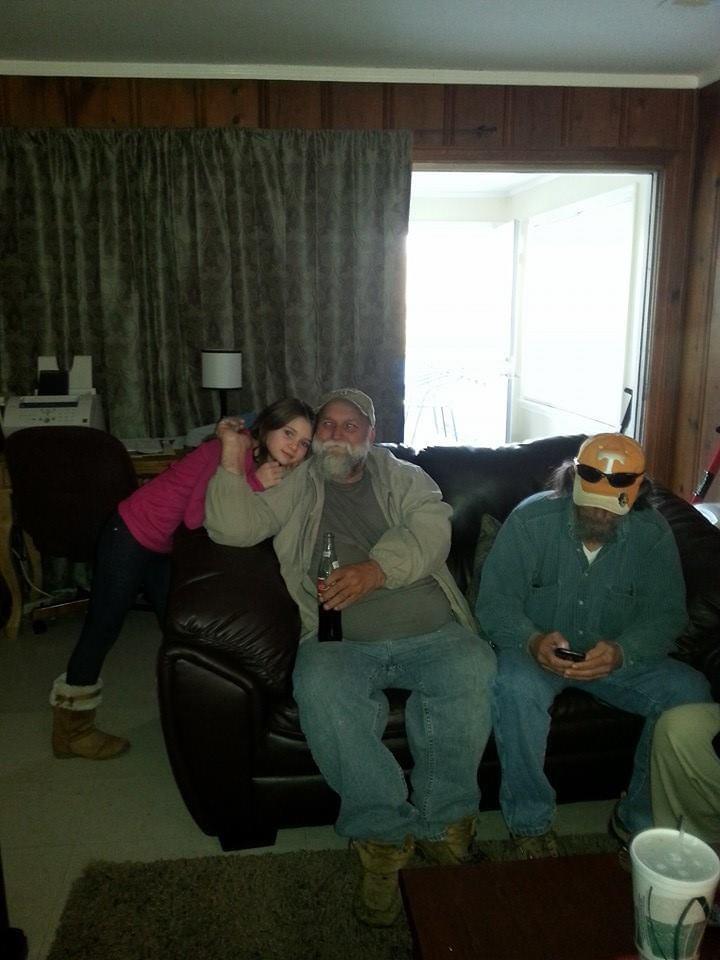 A young girl leans affectionately against a man while another man sits nearby using his phone.