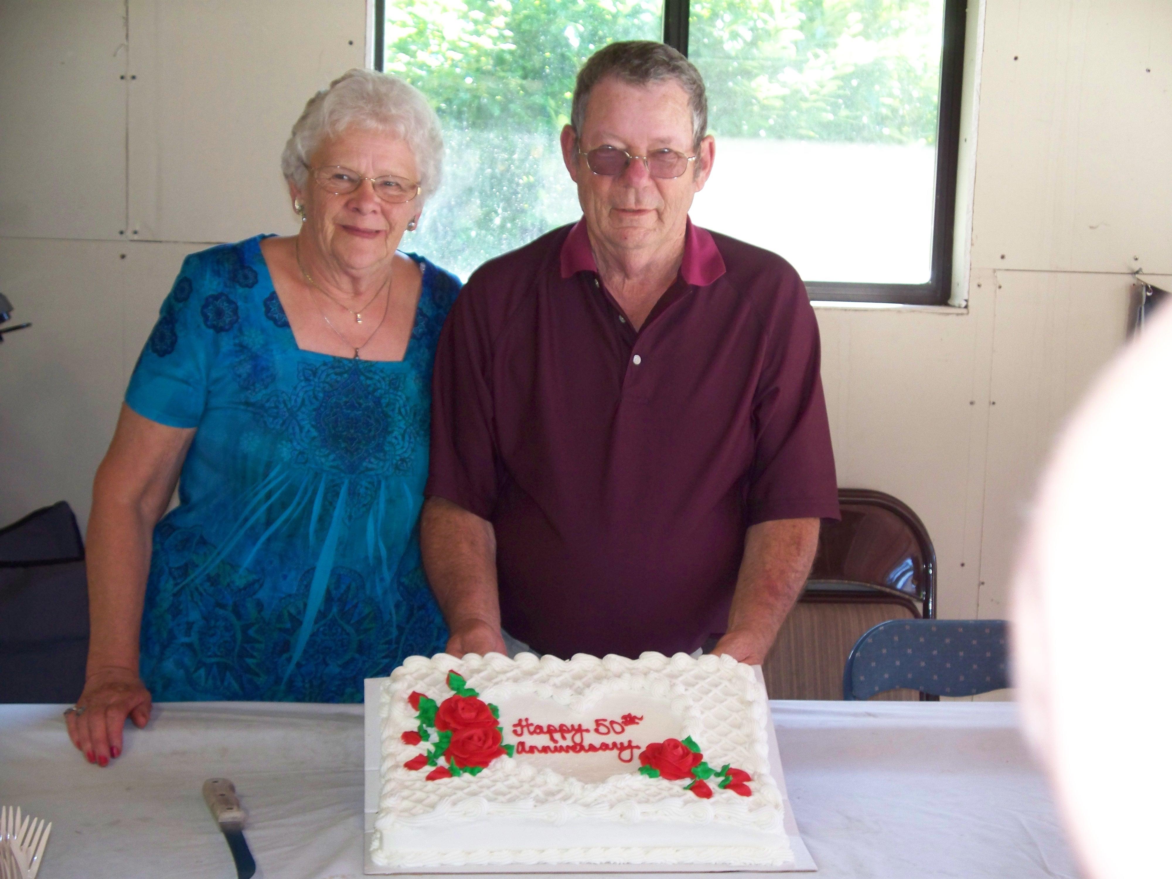 A couple stands proudly beside their anniversary cake adorned with roses at a family celebration.