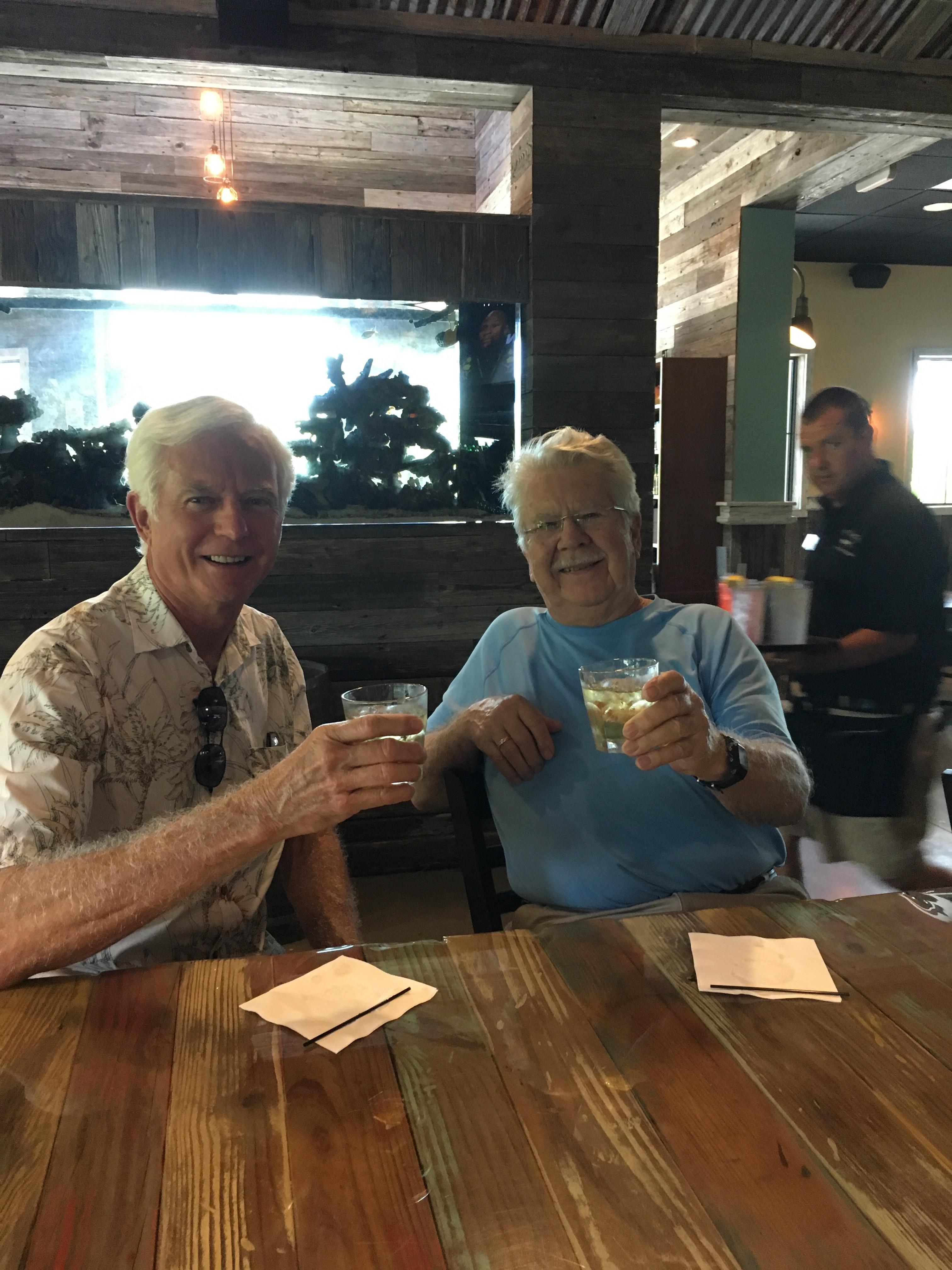 Two older men clink glasses while sitting at a wooden table, smiling joyfully in a warm setting.