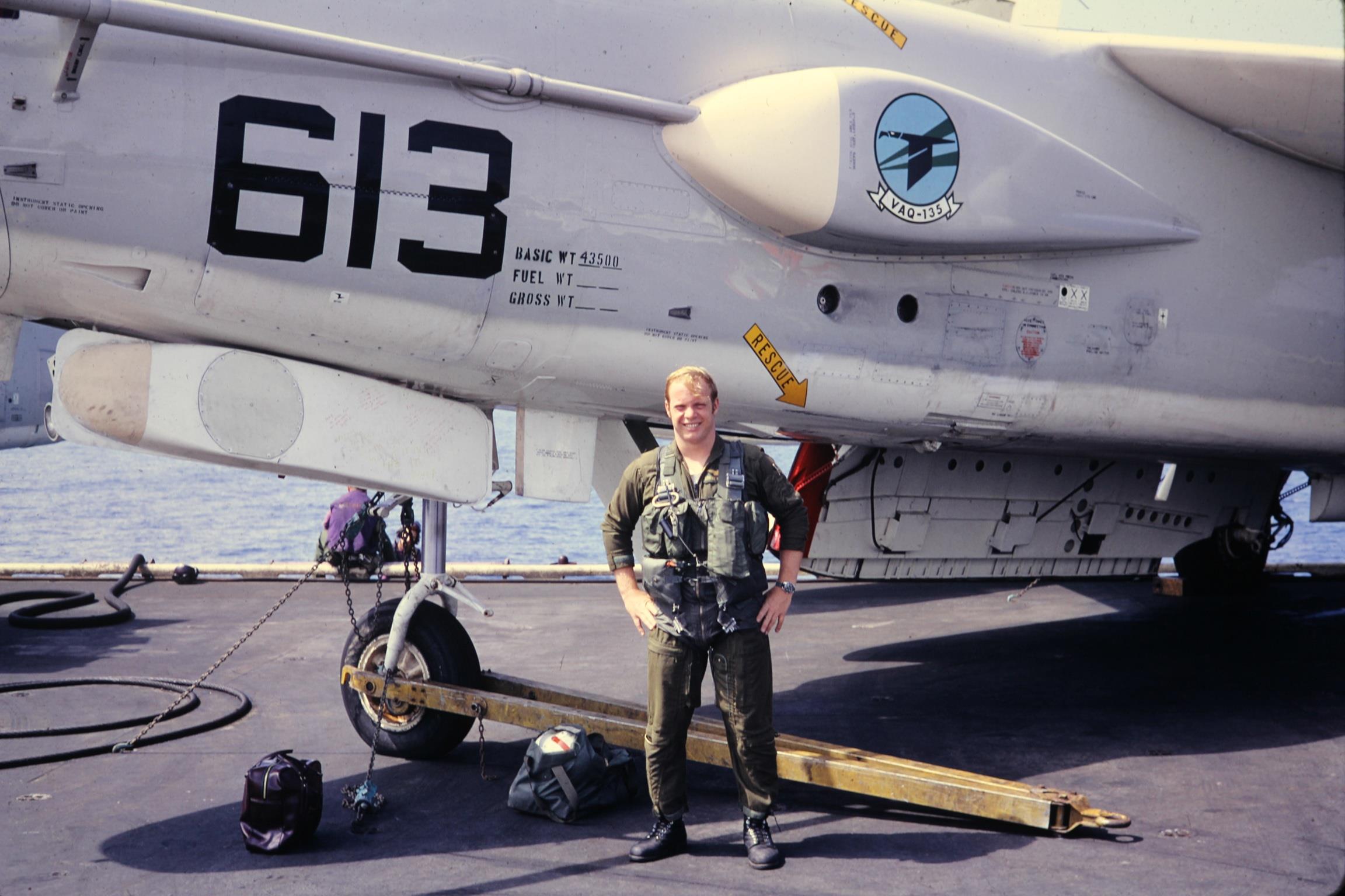 A military pilot poses confidently beside a jet on a carrier deck during bright daylight.