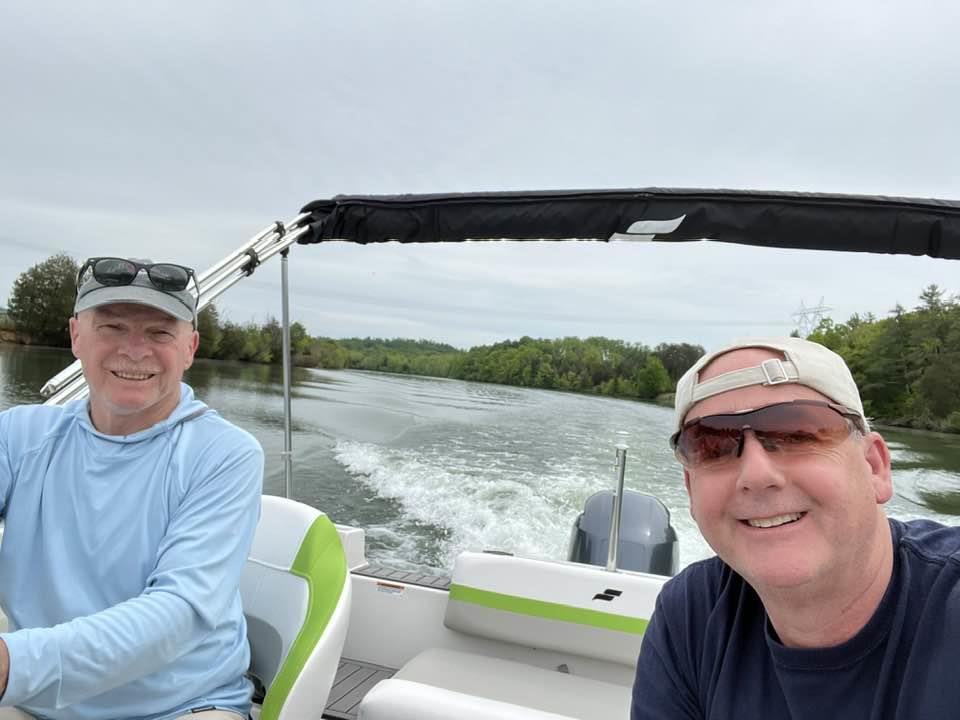 Two men are smiling as they navigate a small boat down a serene river surrounded by greenery.