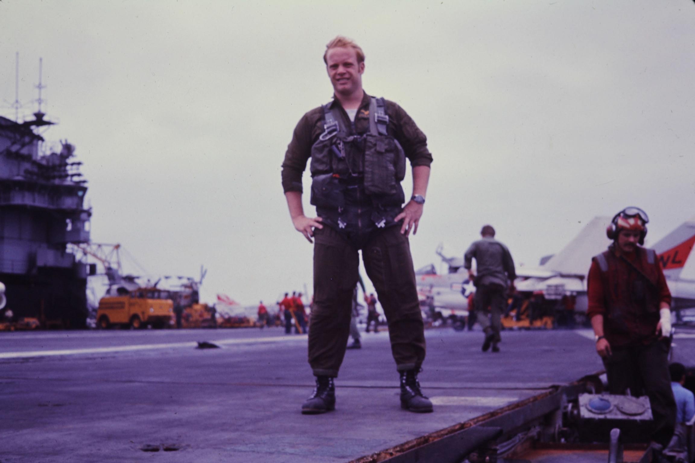Military personnel poses on the deck of an aircraft carrier with planes parked in the background.