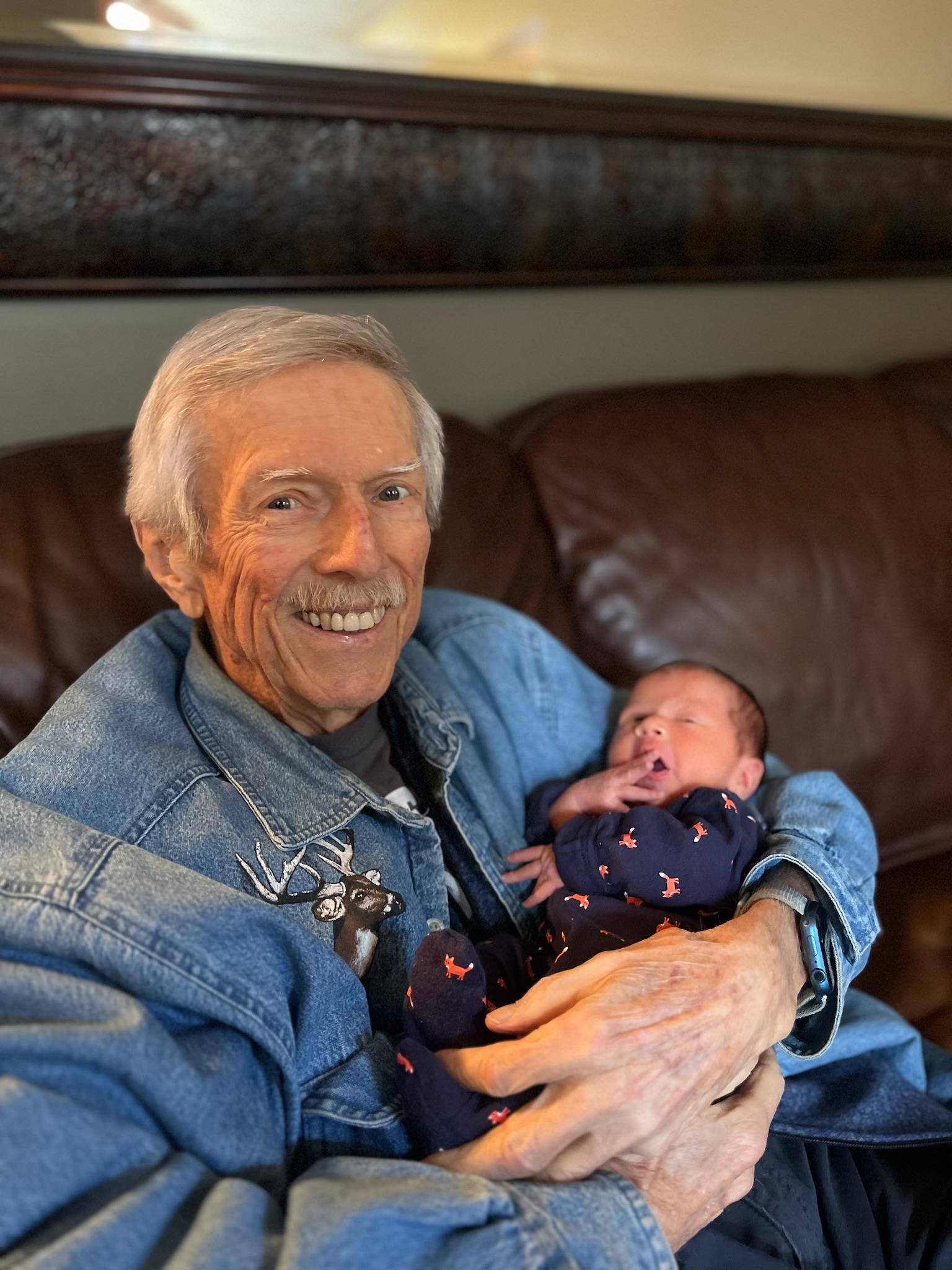 An elderly man smiles, holding a newborn on a leather couch, capturing a warm family moment.