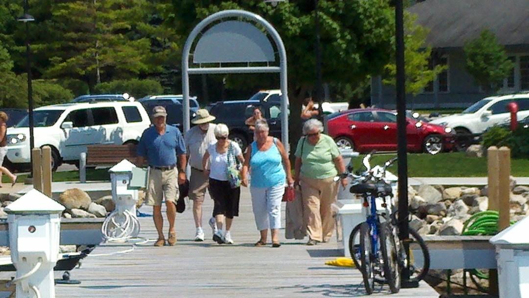 Seniors walk together along a wooden pier enjoying warm weather, surrounded by greenery and boats.