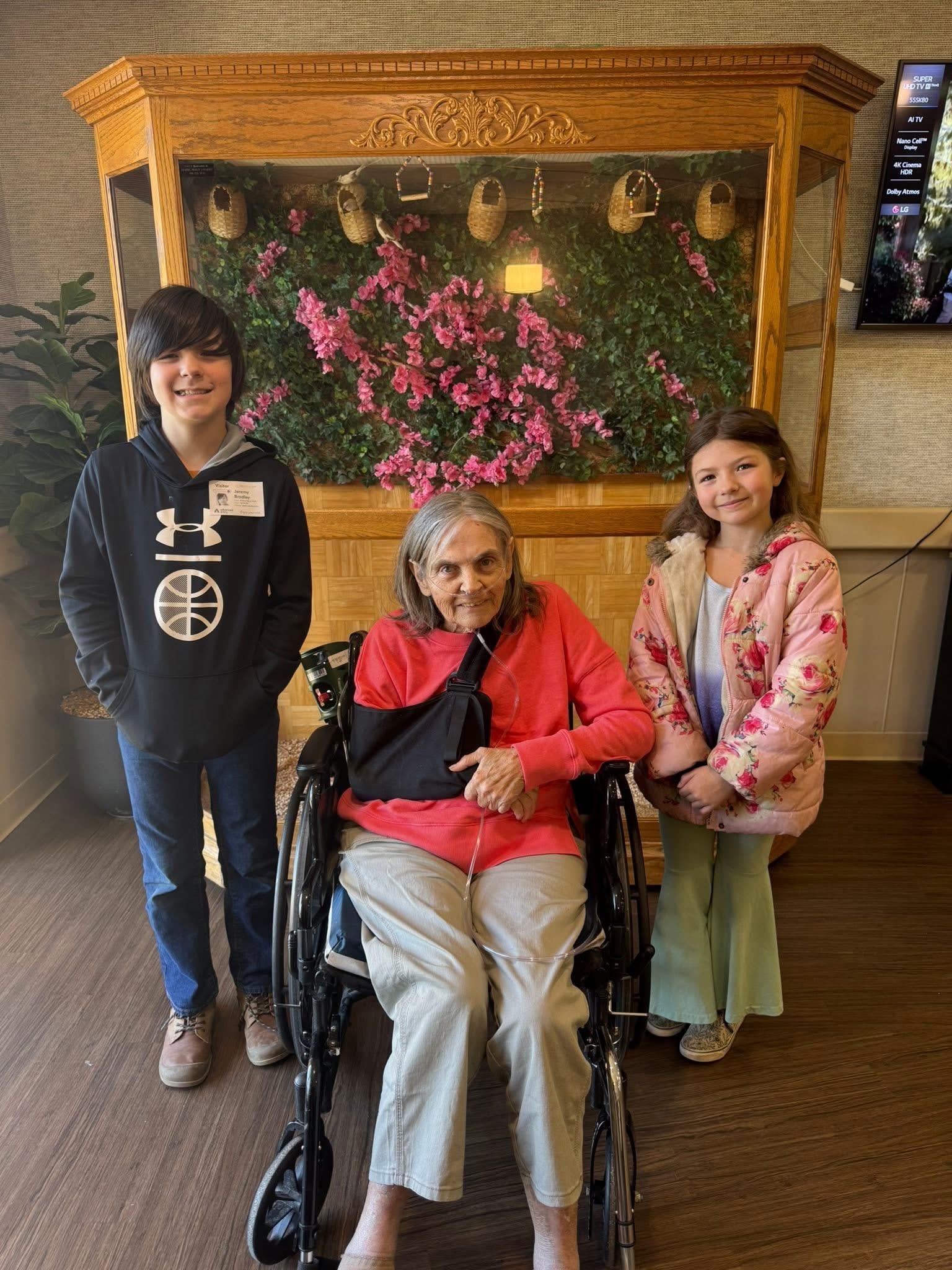 Two children stand beside an elderly woman in a wheelchair, smiling in a nursing home setting.