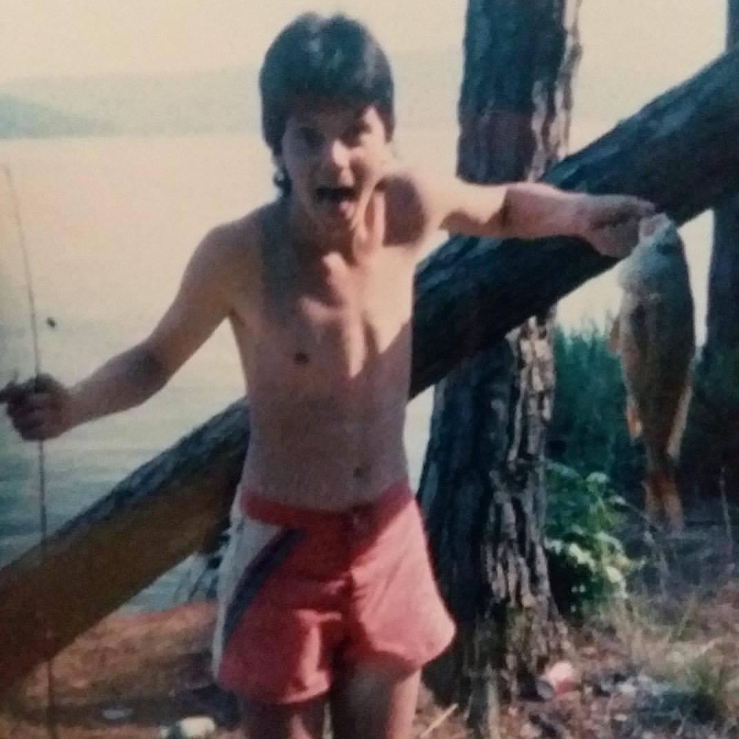 A young boy in shorts shows excitement holding a large fish near a lake on a sunny day.