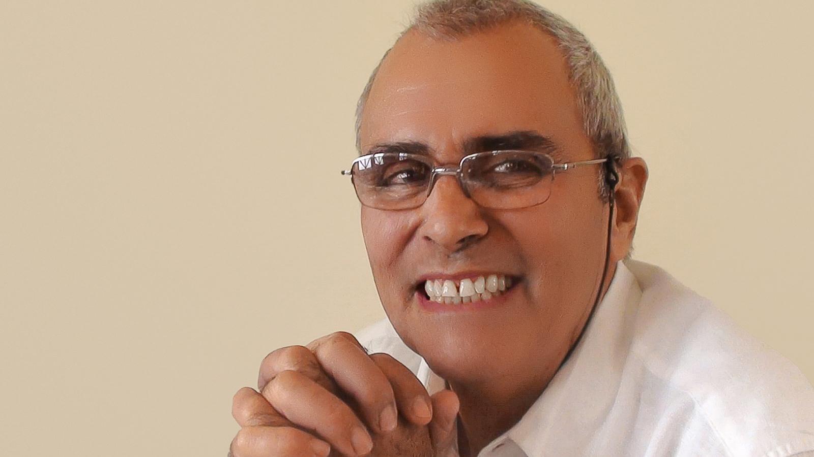 A cheerful elderly man with glasses smiles widely in a cozy, warm setting.