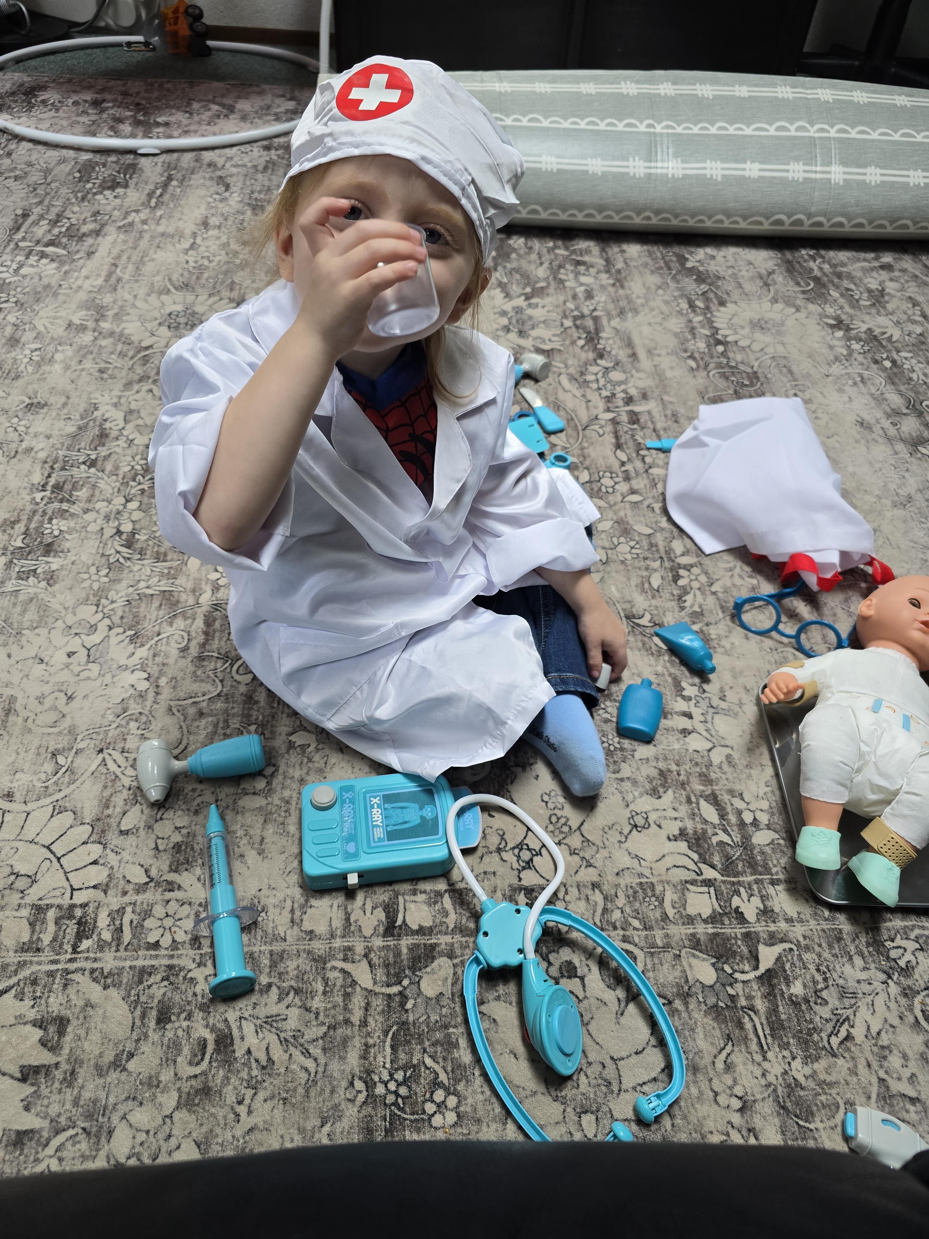 A young child wearing a doctor's coat drinks from a cup while surrounded by toys and medical tools.