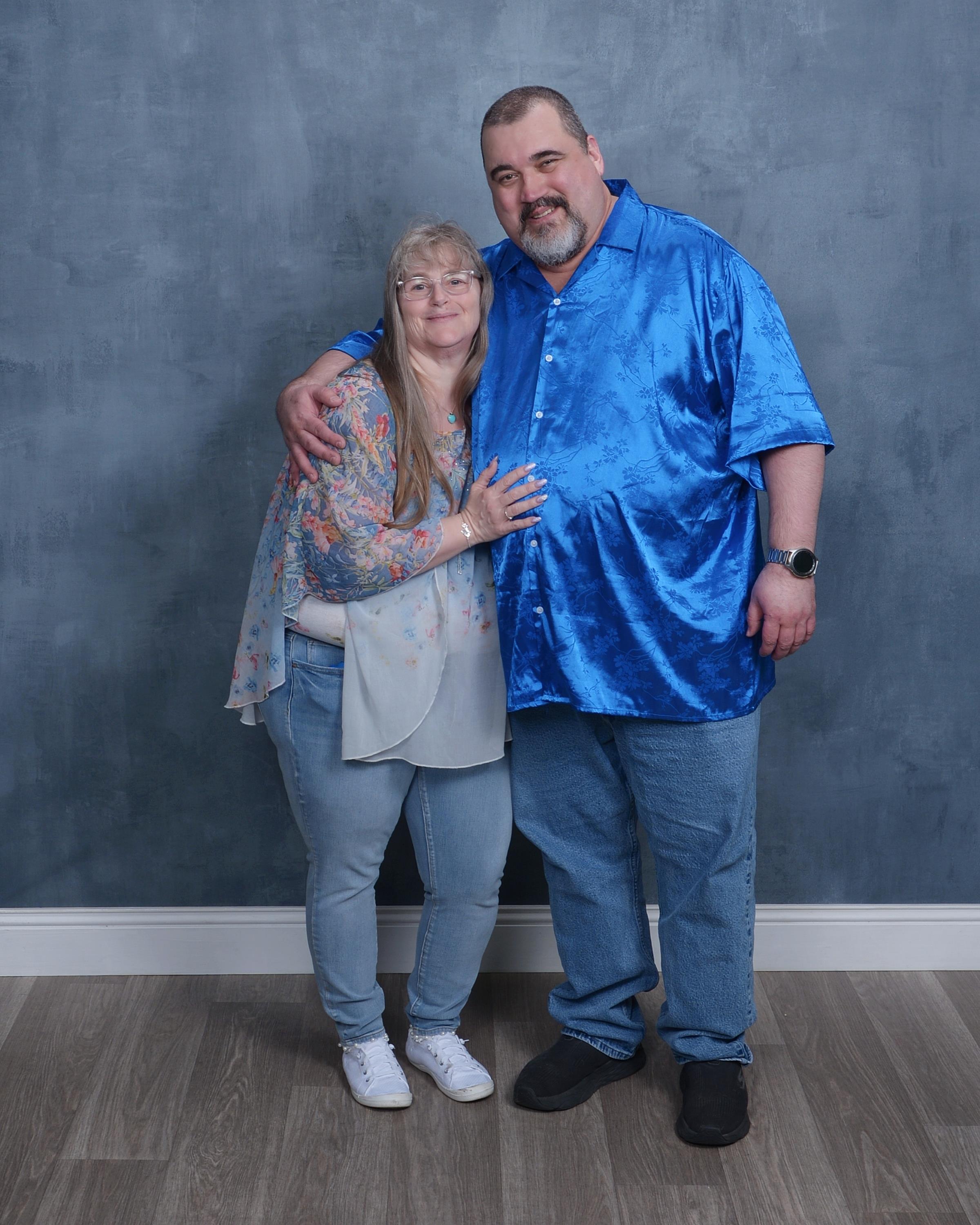 A couple stands closely together, smiling warmly against a blue background in a studio setting.