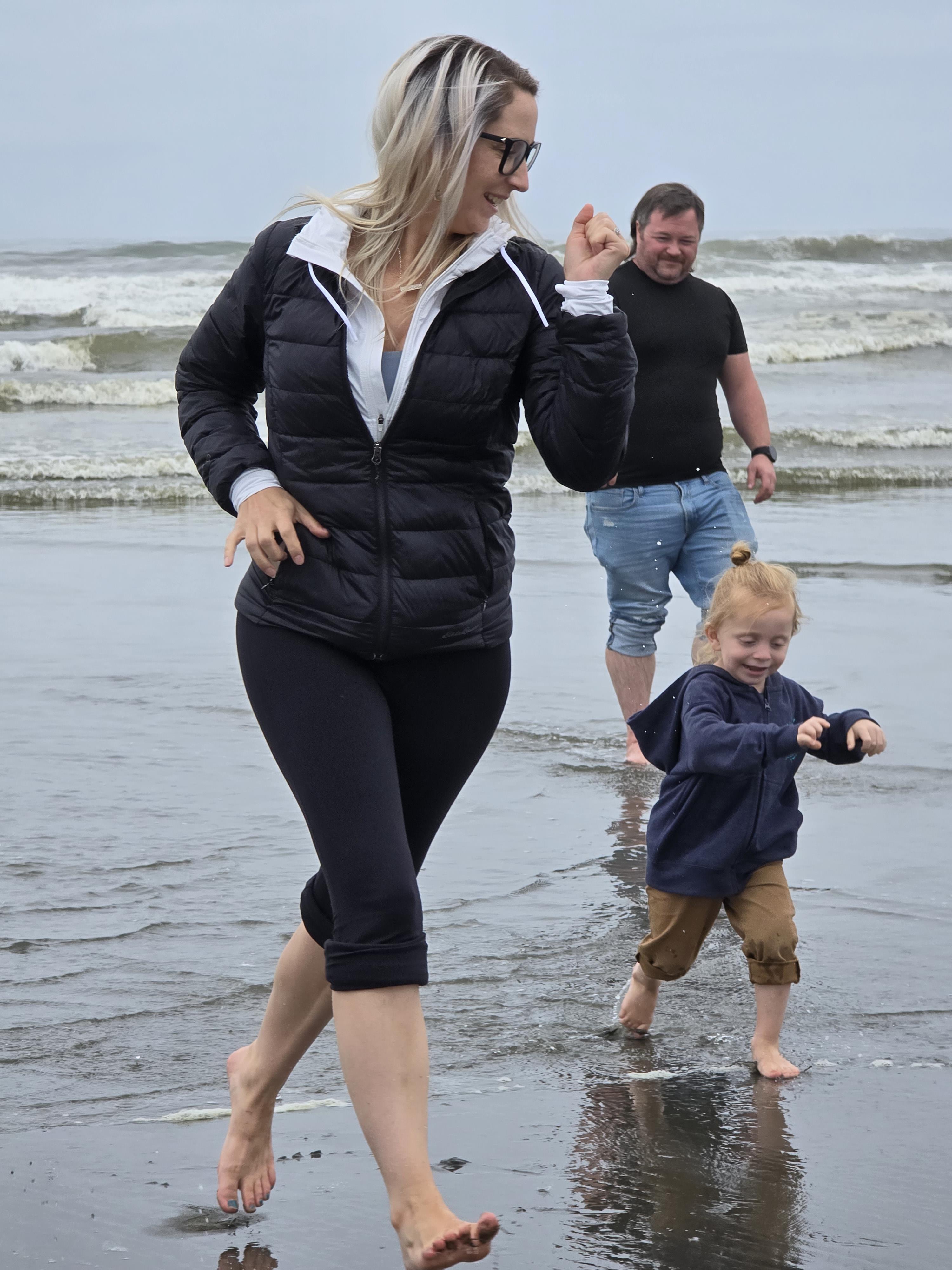 A woman and her child play joyfully in the shallow water at the beach while a man watches.