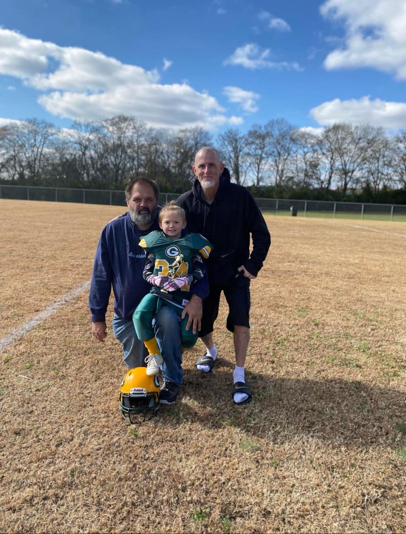Two men pose with a young boy in a football uniform, smiling on a grassy field under a clear sky.