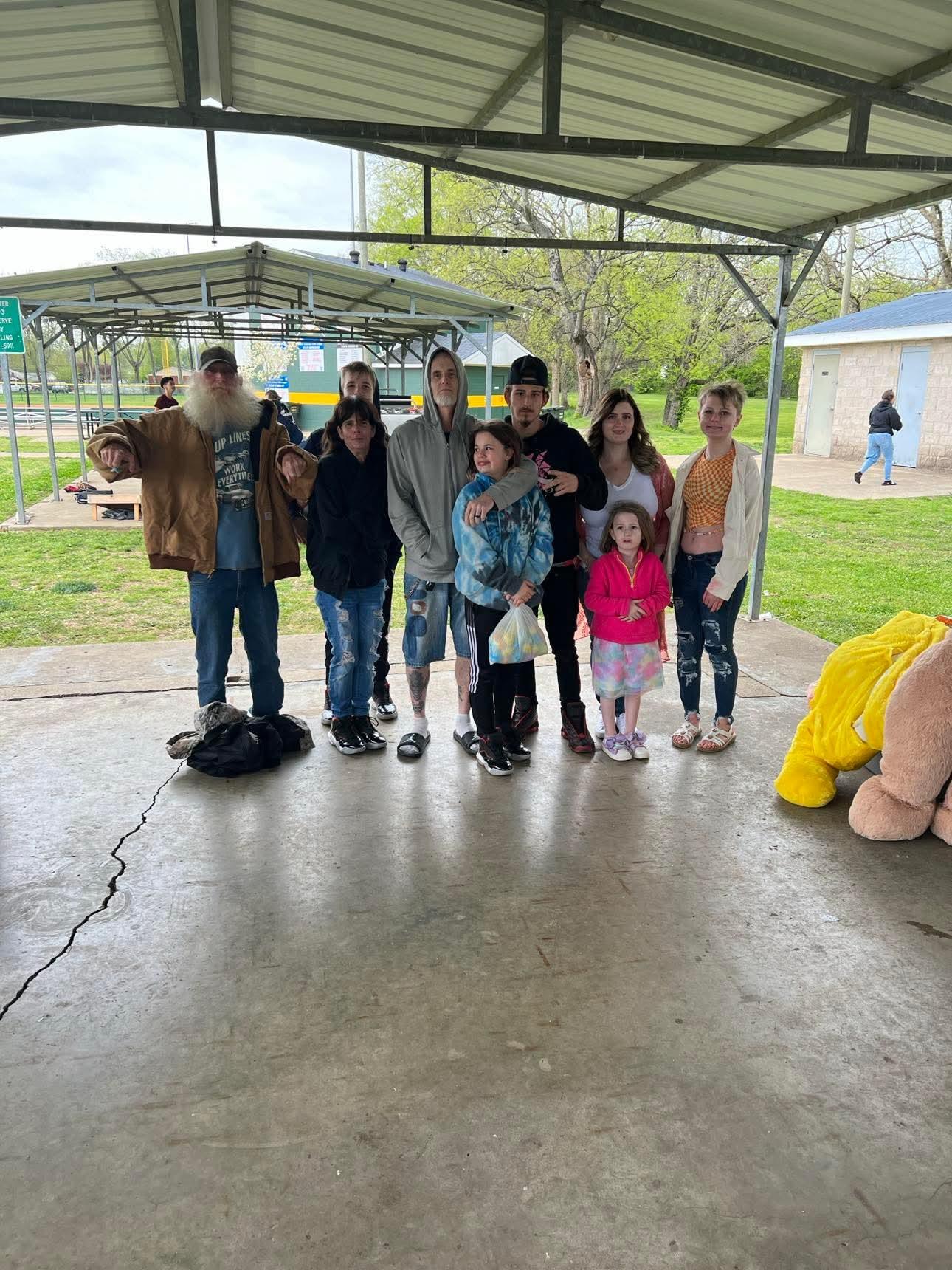 A family stands together under a shelter in a park on a cloudy spring day, smiling happily.