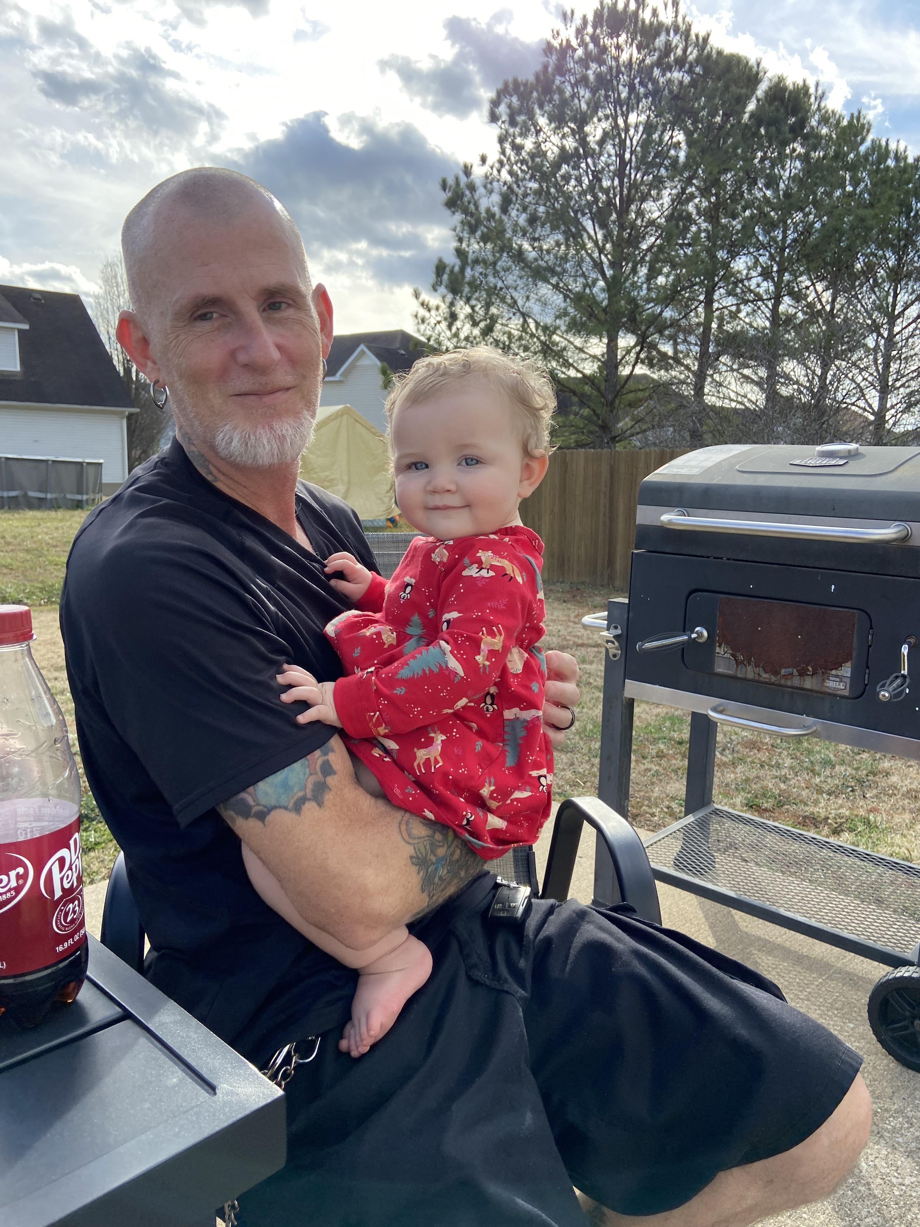 A man enjoys quality time with a toddler on his lap while grilling outdoors on a cloudy day.