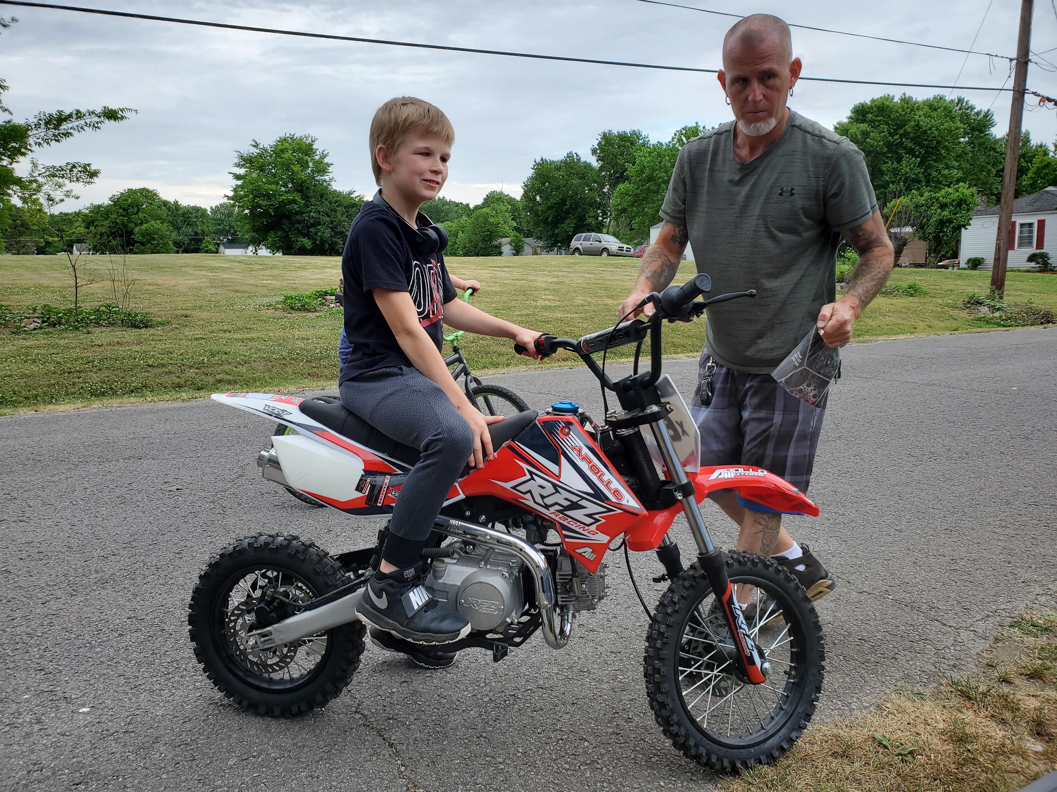 A young boy sits on a dirt bike while an adult stands beside him on a sunny day.
