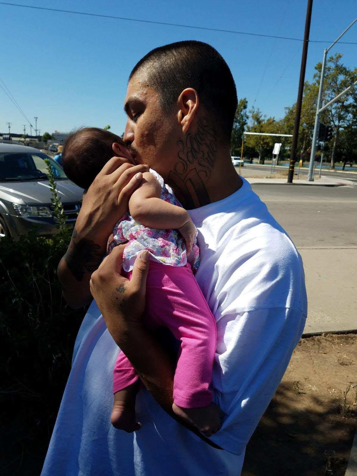 A man lovingly kisses his baby daughter while holding her on a warm, clear day.
