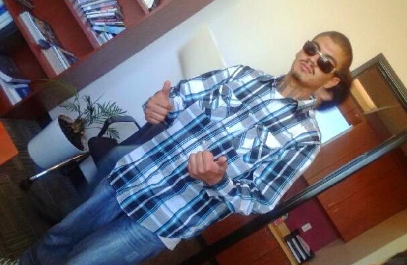 A person stands casually in a room, wearing sunglasses and a plaid shirt, giving a thumbs-up.