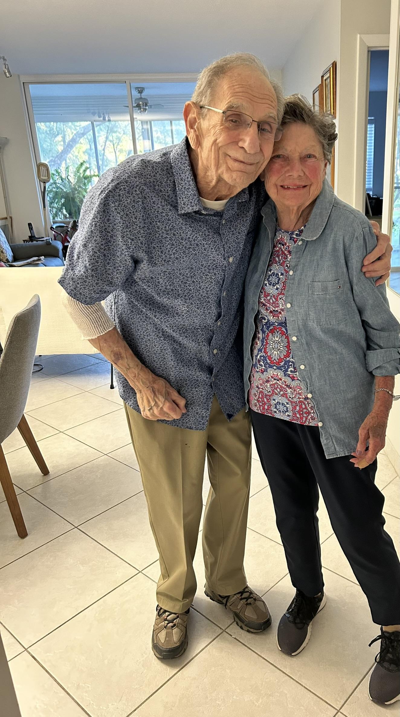 Couple stands close together, smiling warmly in a well-lit living room filled with plants.