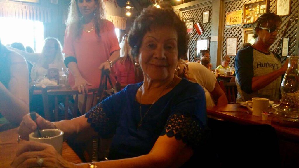 An elderly woman with a warm smile sips coffee at a bustling cafe filled with patrons.