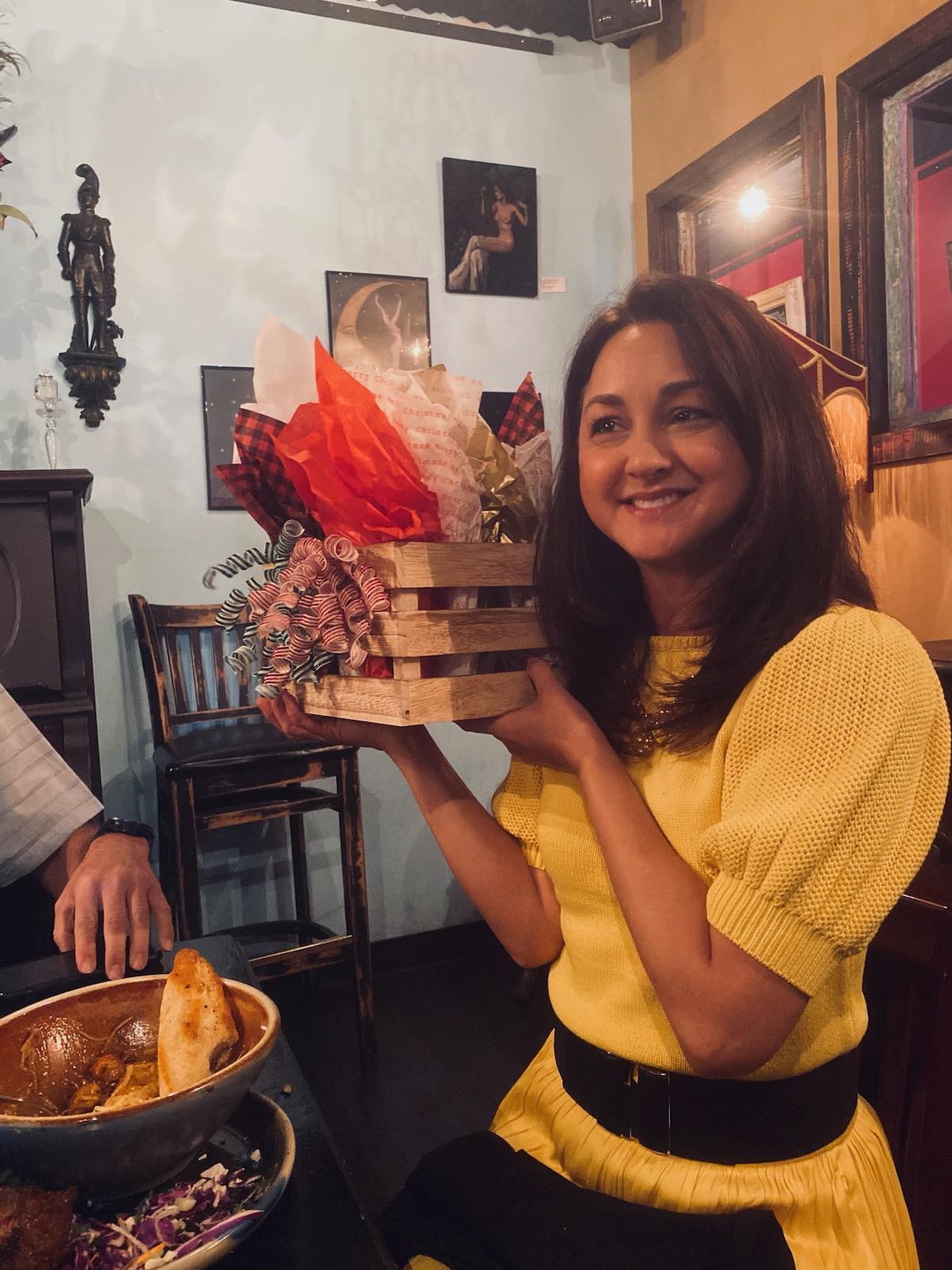 A woman joyfully holds a beautifully arranged gift basket while dining with friends.