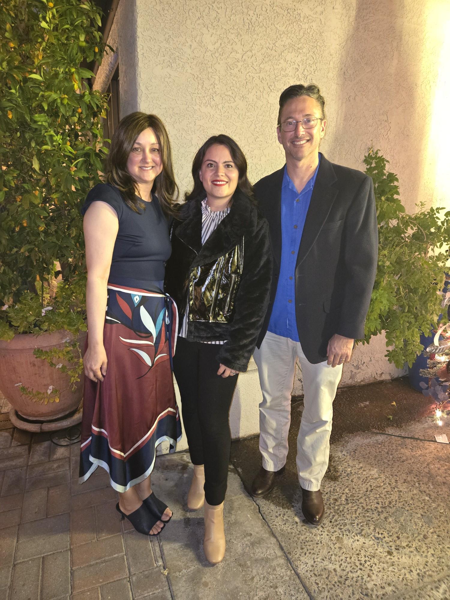 Three friends smile and pose for a picture during a social event at night near a wall.
