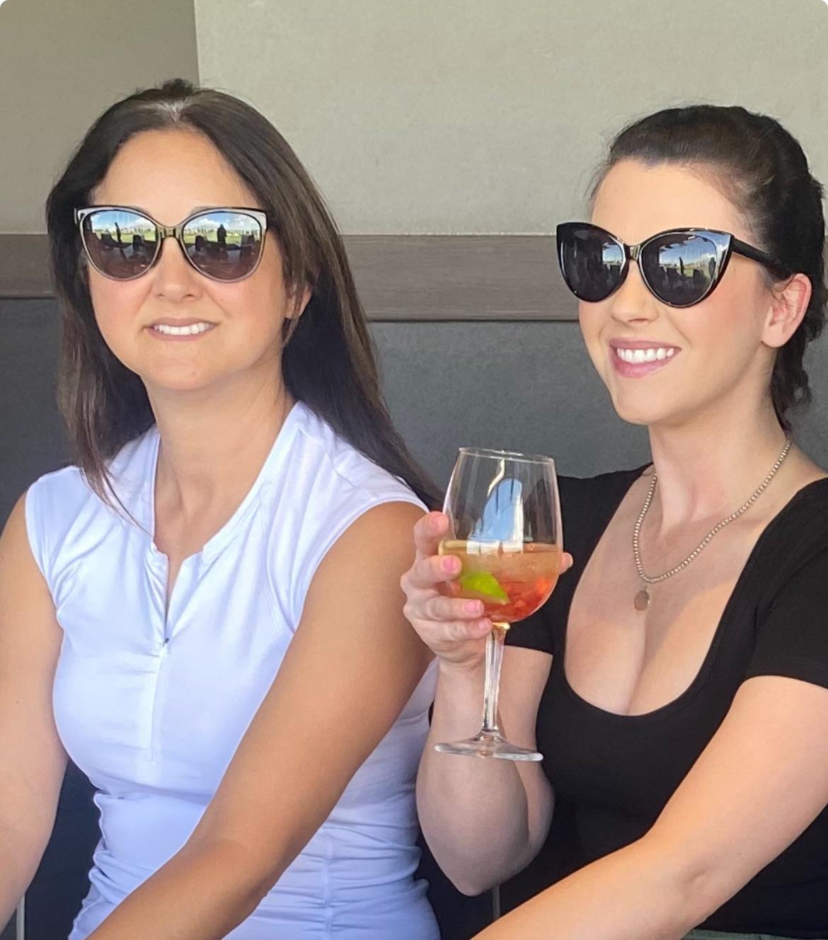 Two friends sit together, one holding a cocktail, enjoying a sunny day outdoors.