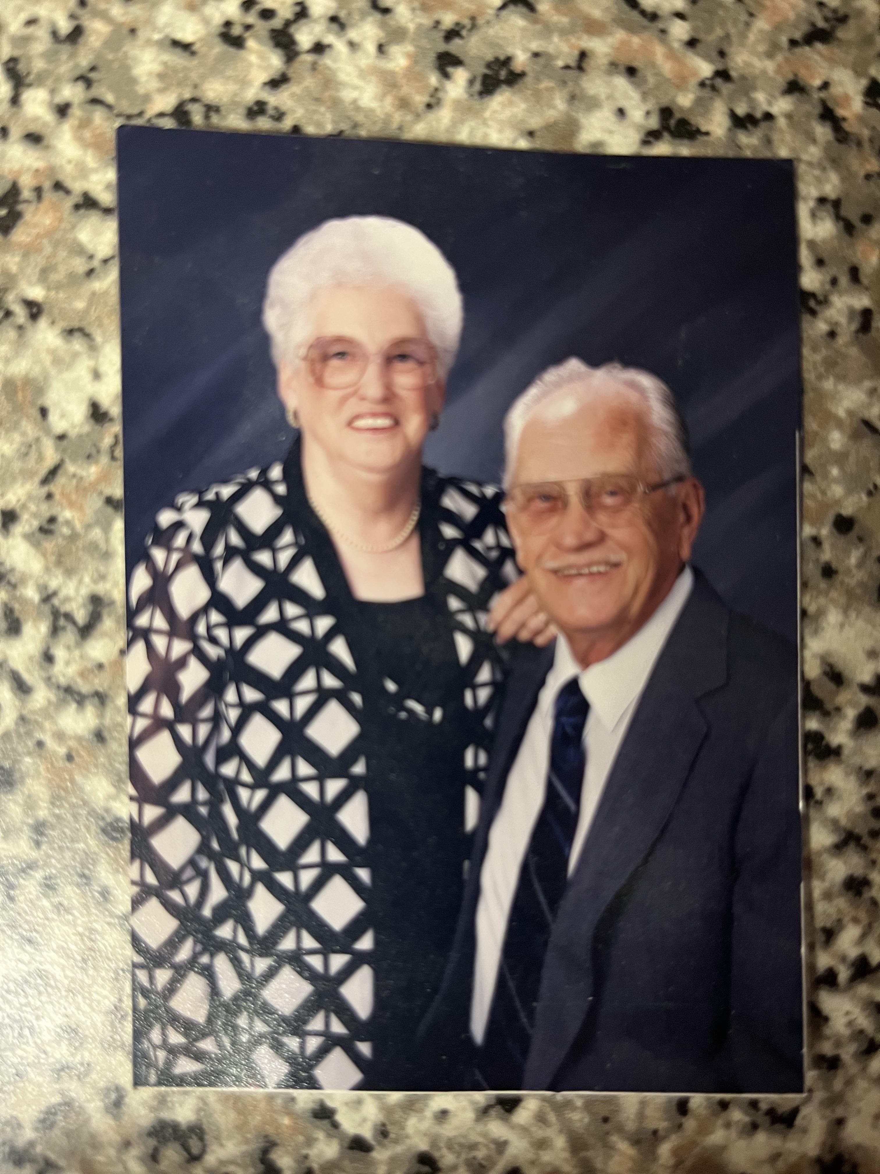 An elderly couple stands close, dressed in formal outfits, smiling happily at the camera.