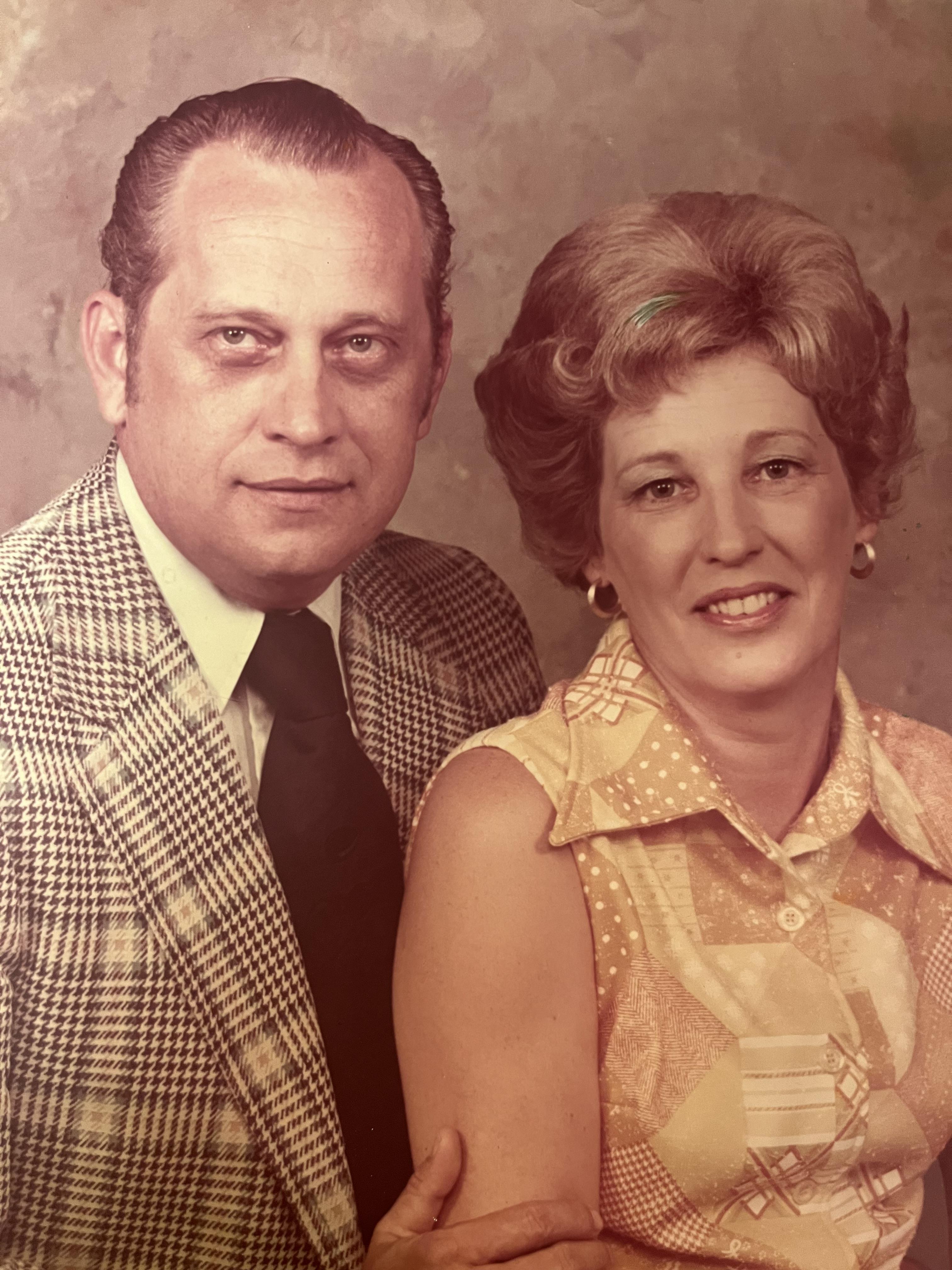 Elderly couple stands close together, smiling warmly in a vintage-styled studio setting.