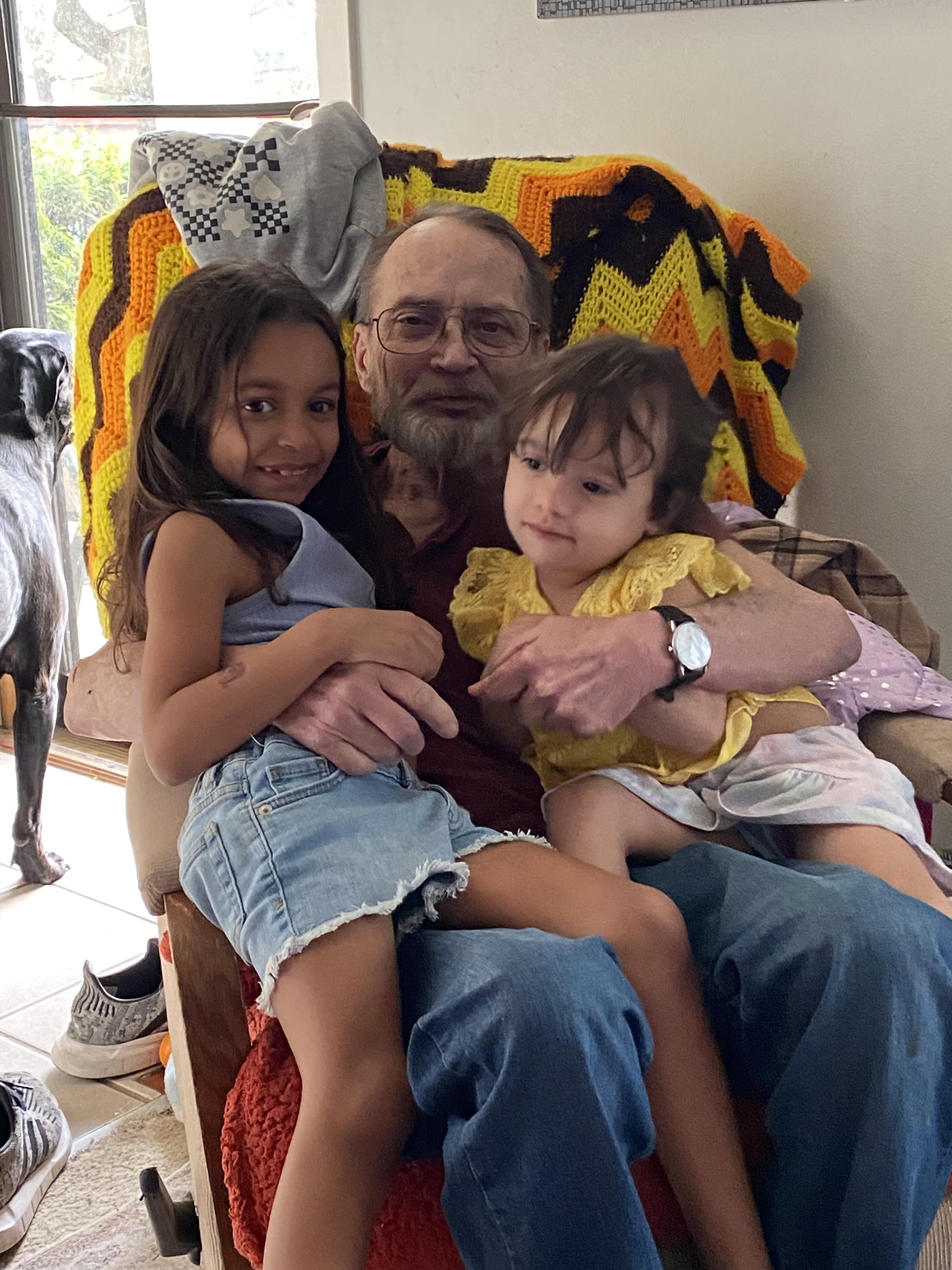 Grandfather smiles as he holds his two grandchildren on his lap in a warm living room.