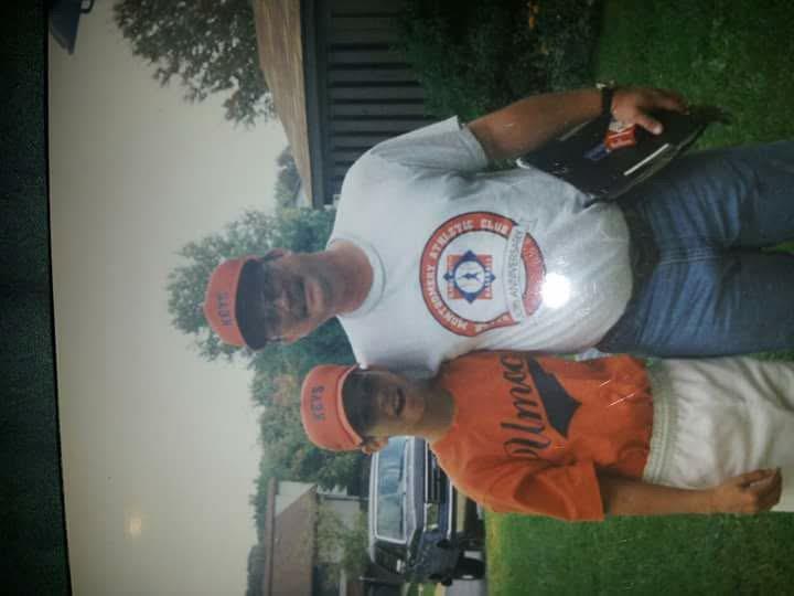 A father and son pose together outdoors wearing matching caps, smiling happily under a cloudy sky.