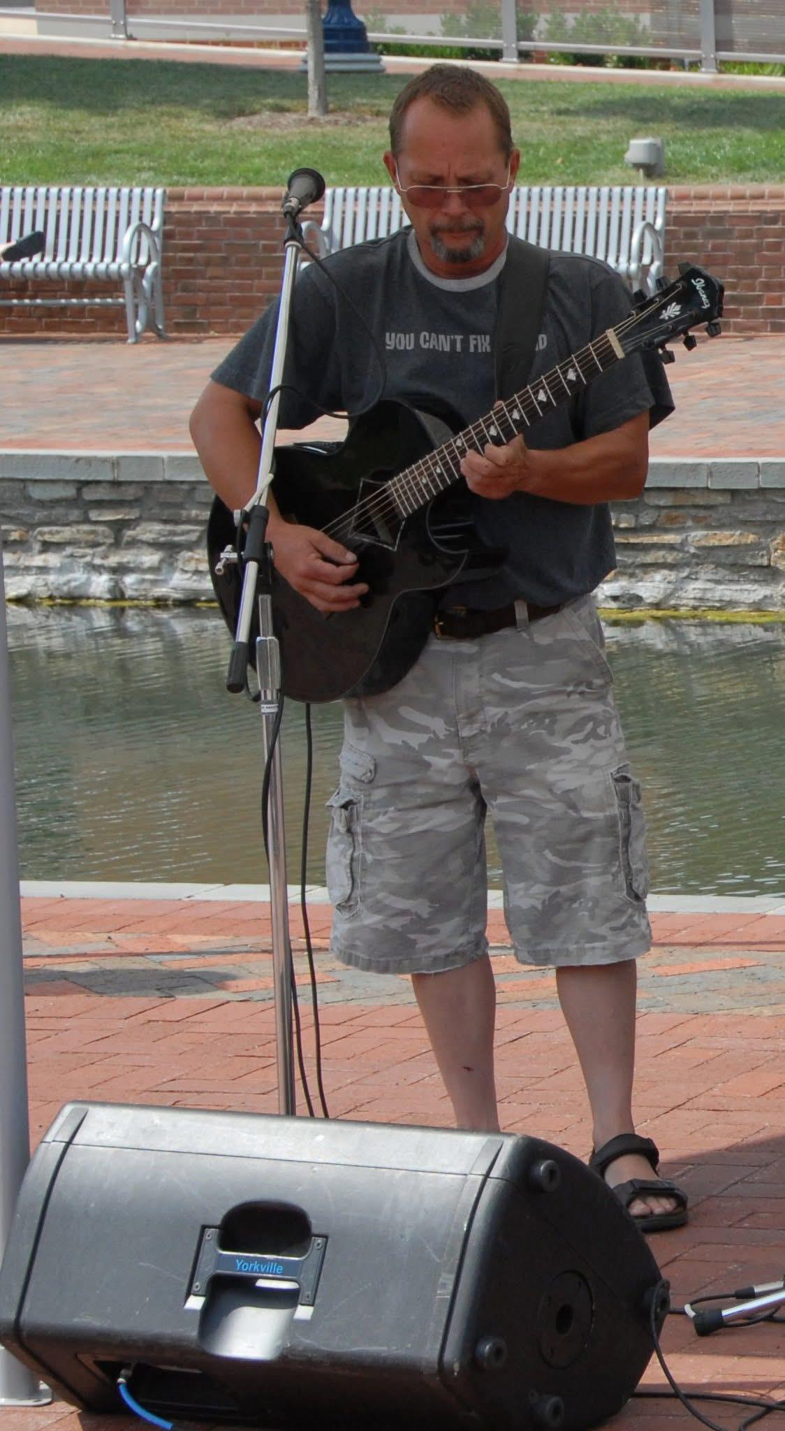 A musician plays acoustic guitar while standing near the water in a lively park setting.