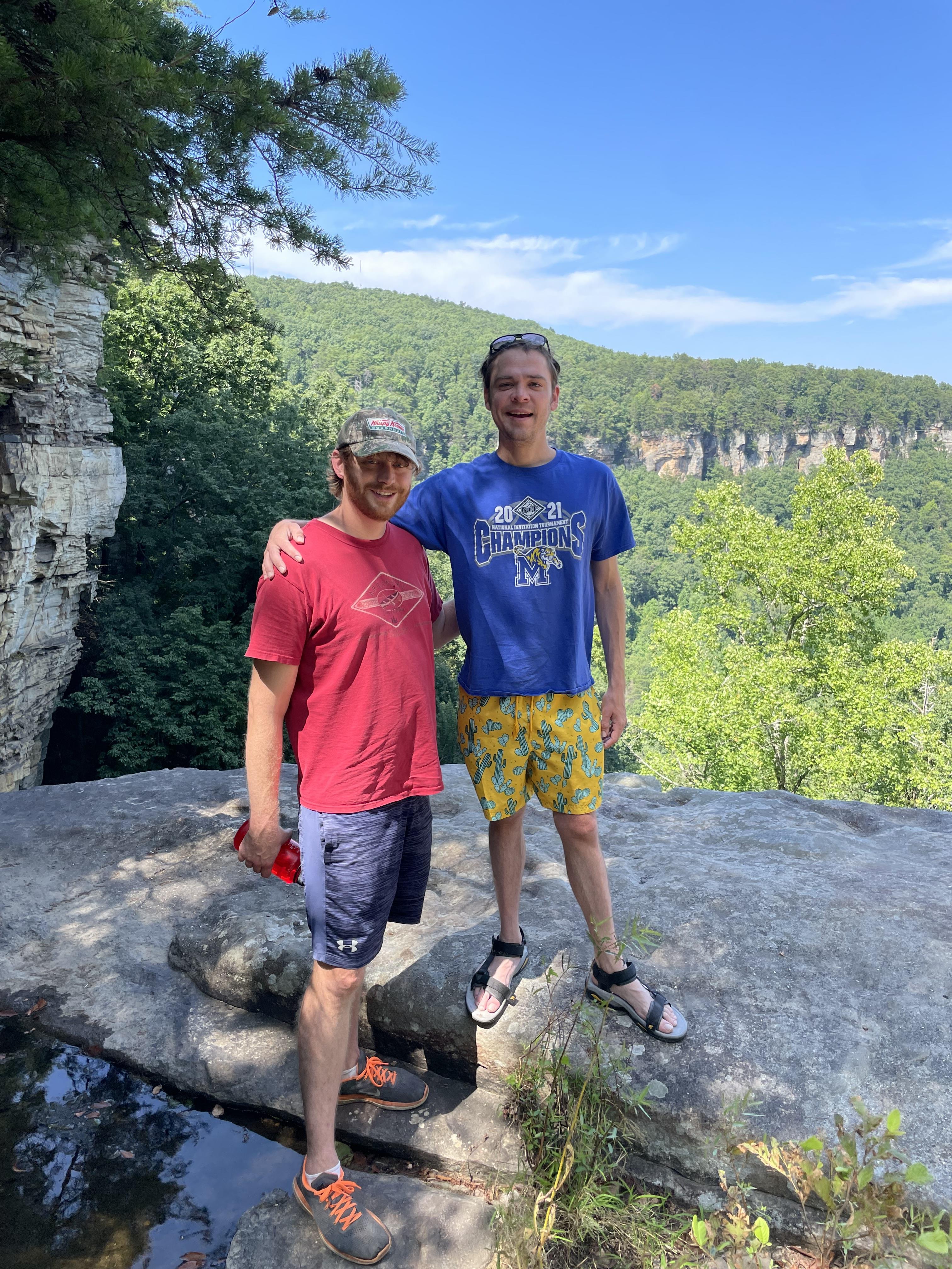 Two friends stand on a rocky ledge, smiling amid lush trees and a clear blue sky.