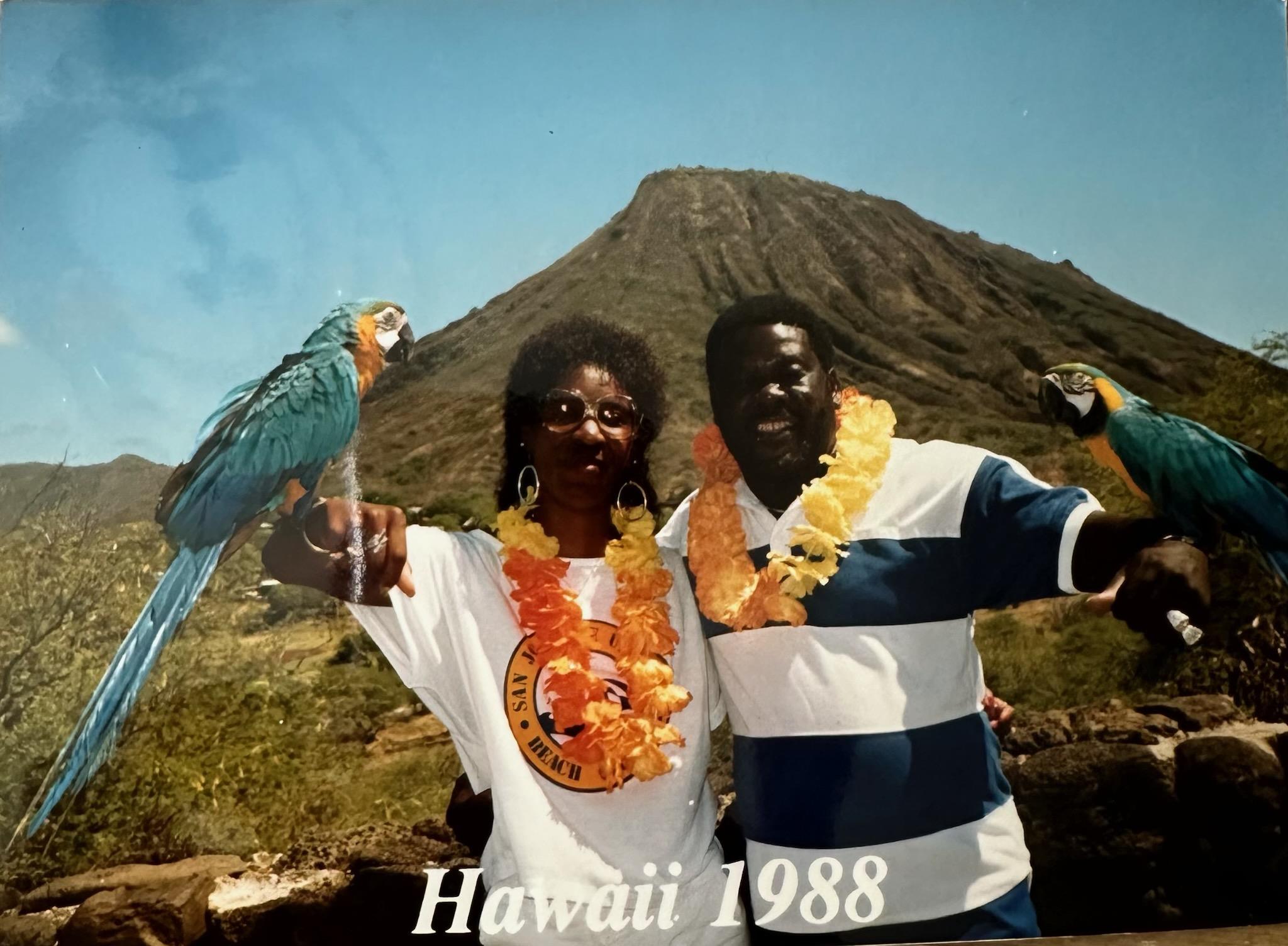 Two people wearing leis and smiling with parrots at a volcanic location in Hawaii.