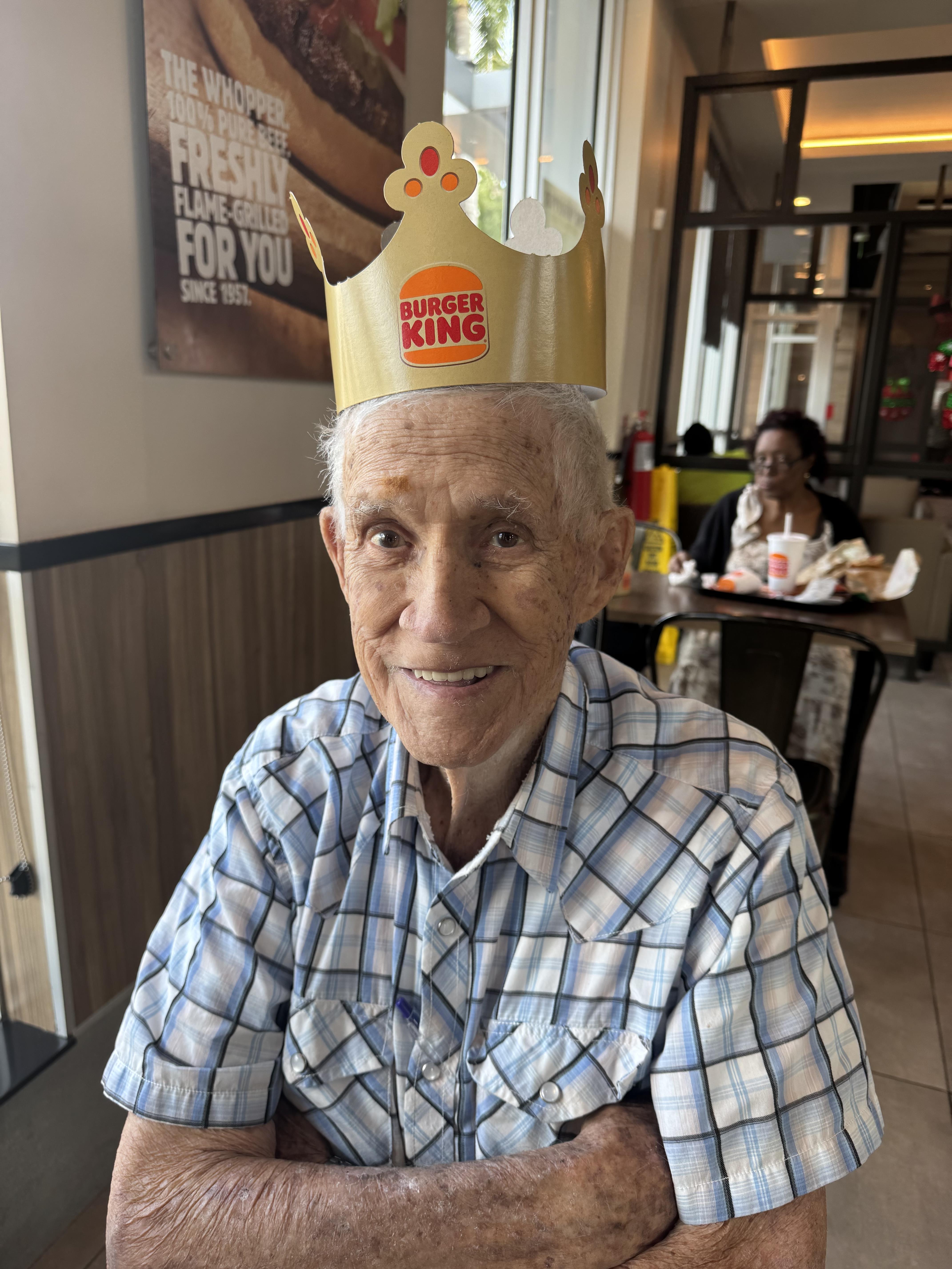 A cheerful elderly man dons a colorful crown while enjoying a meal in a lively dining place.