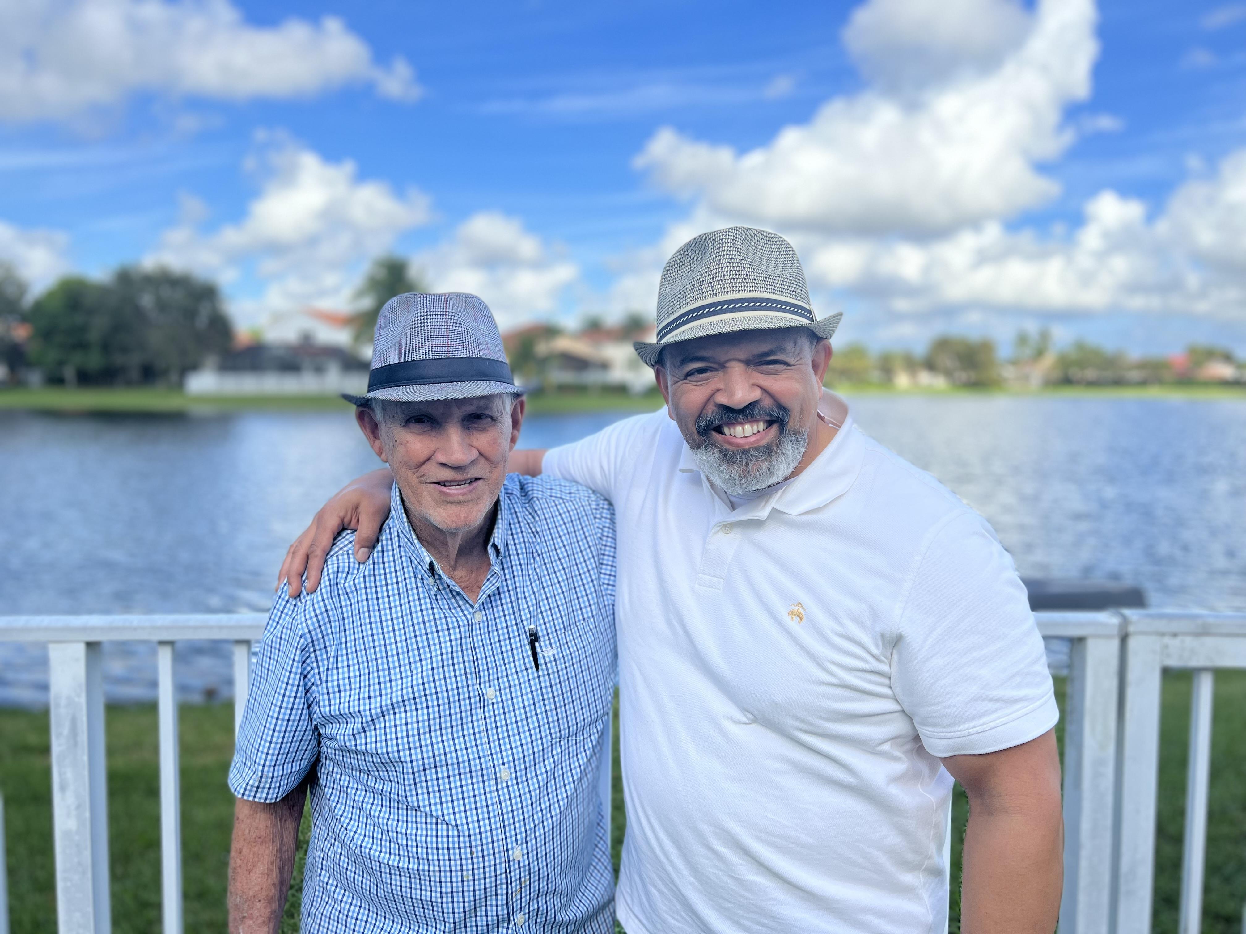 Two men enjoy each other's company near a calm lake, wearing matching hats and smiling broadly.