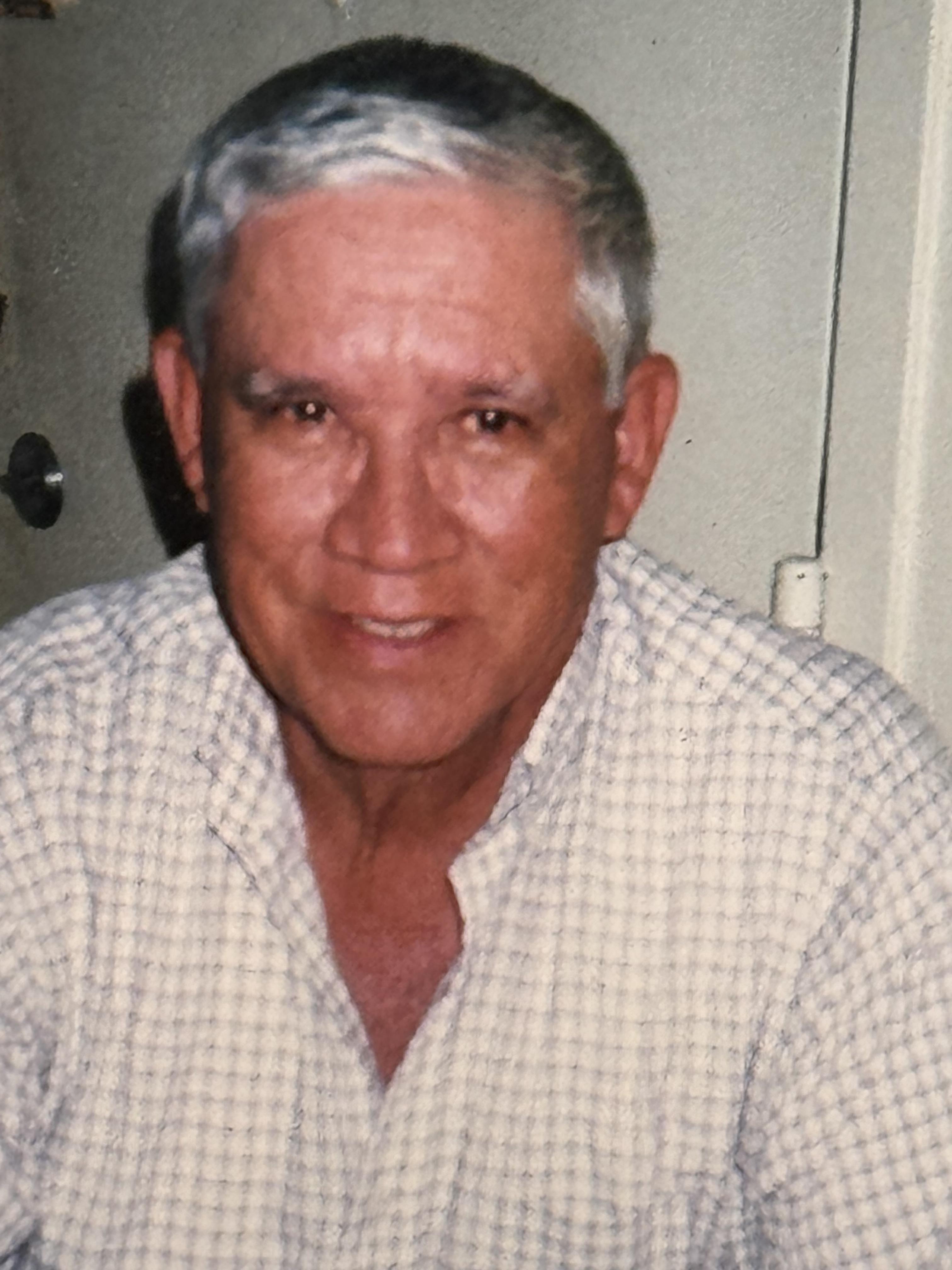 An elderly man with gray hair is smiling gently at the camera while seated indoors.