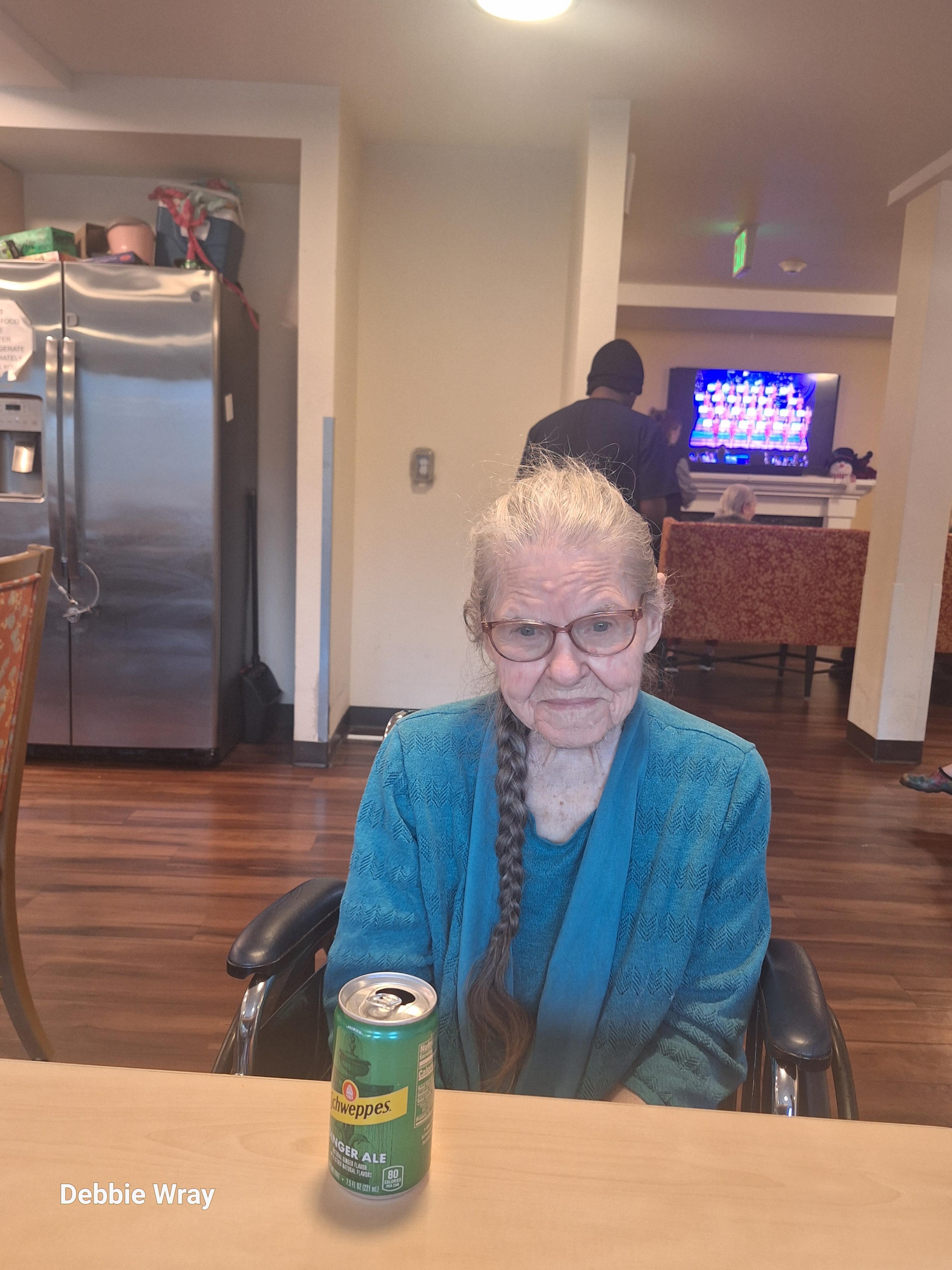 An elderly woman sits in a wheelchair with a can of soda beside her, watching TV in a lively room.