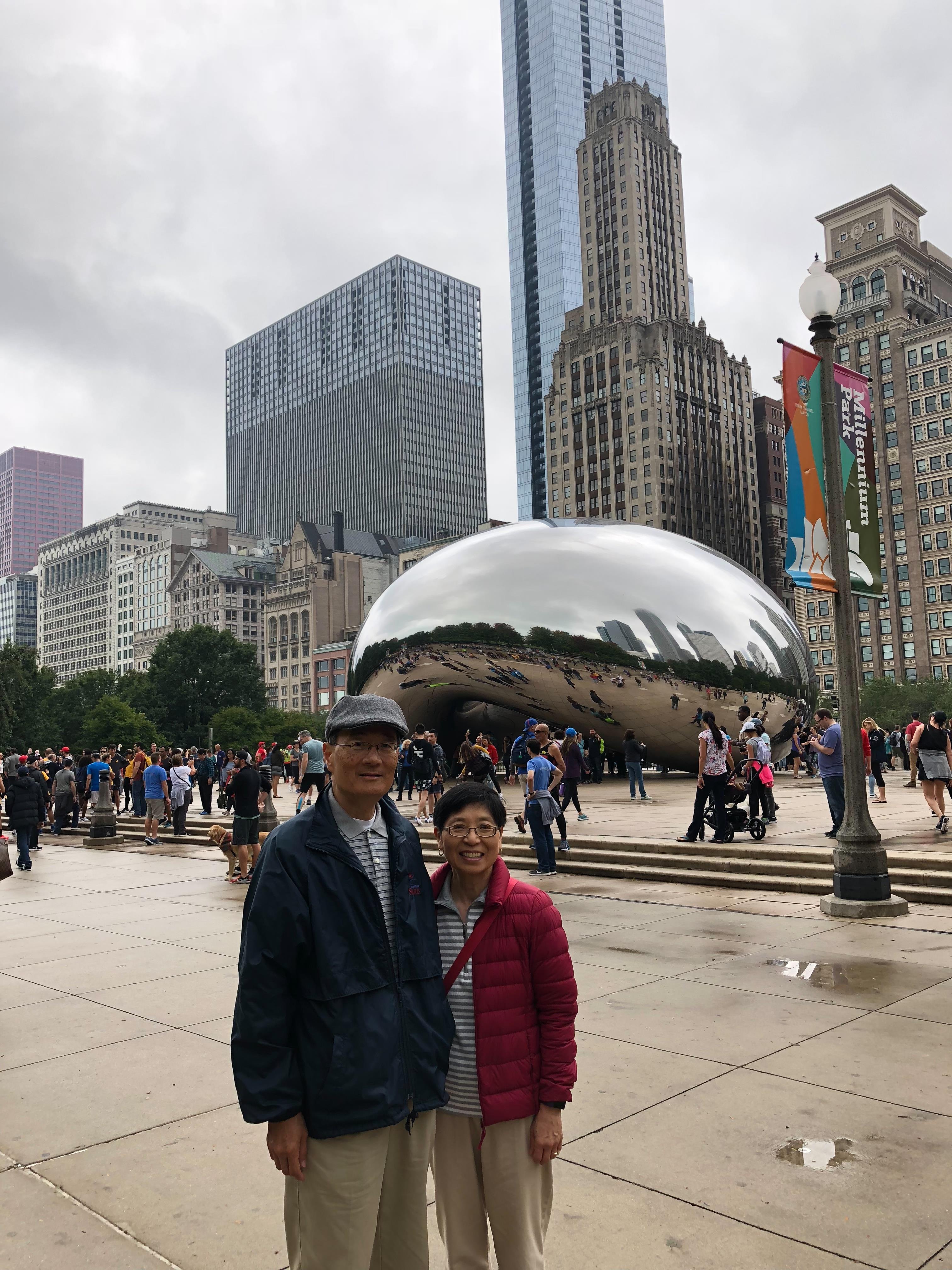 Two people smile by a large, shiny sculpture among city buildings and visitors.