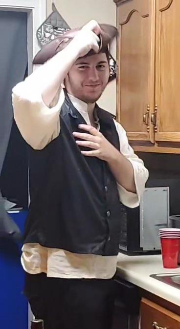 A man dressed in period clothing gestures playfully in a cozy kitchen filled with friends.