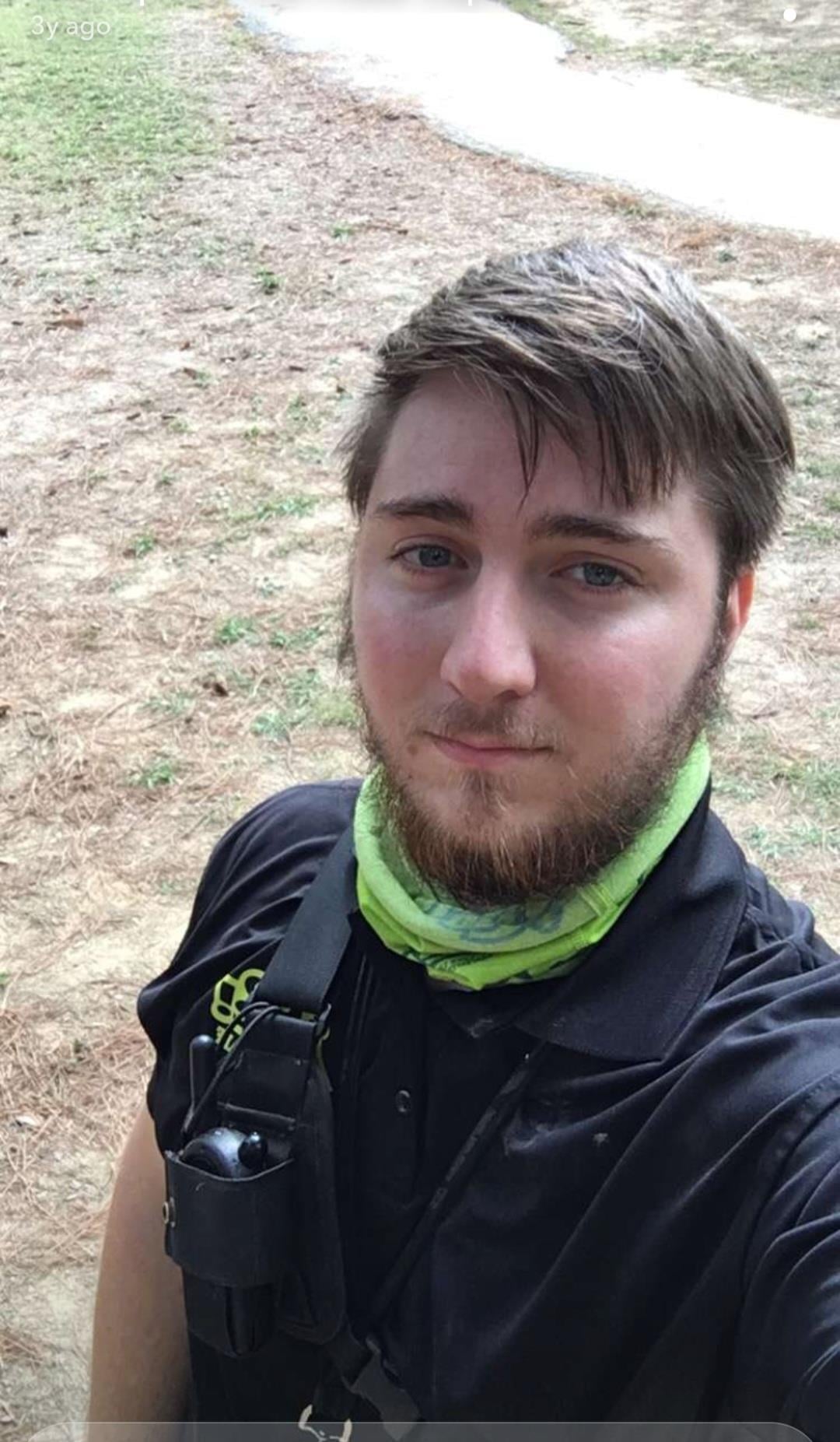 A young man outdoors, smiling in casual clothes and a green scarf.