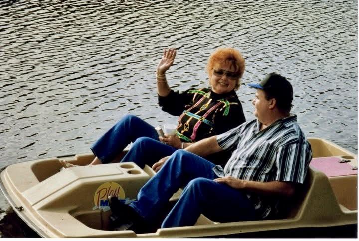 Two people relax on a pedal boat, smiling and waving, surrounded by calm water and greenery.
