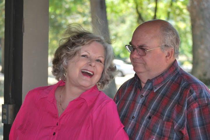 A woman in a pink shirt smiles joyfully while a man in plaid looks on with a neutral expression.